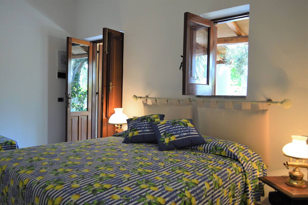 A bedroom with a bed, two pillows, a nightstand with a lamp, a window with an open door, and a view of greenery outside.