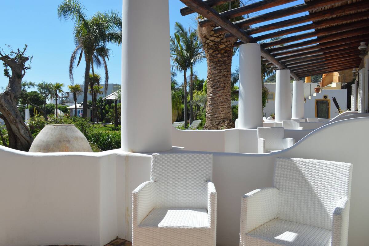 A white wicker chair under a wooden pergola with a view of palm trees and a white building in the background.