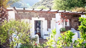 A stone building with a wooden pergola, white furniture, and a garden with flowering plants. - Hotel Oasi (Lipari)