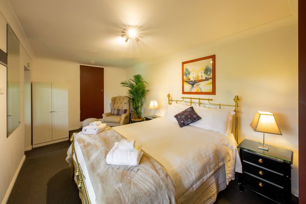 A hotel room with a large bed, a chair, a nightstand with a lamp, and a painting on the wall.