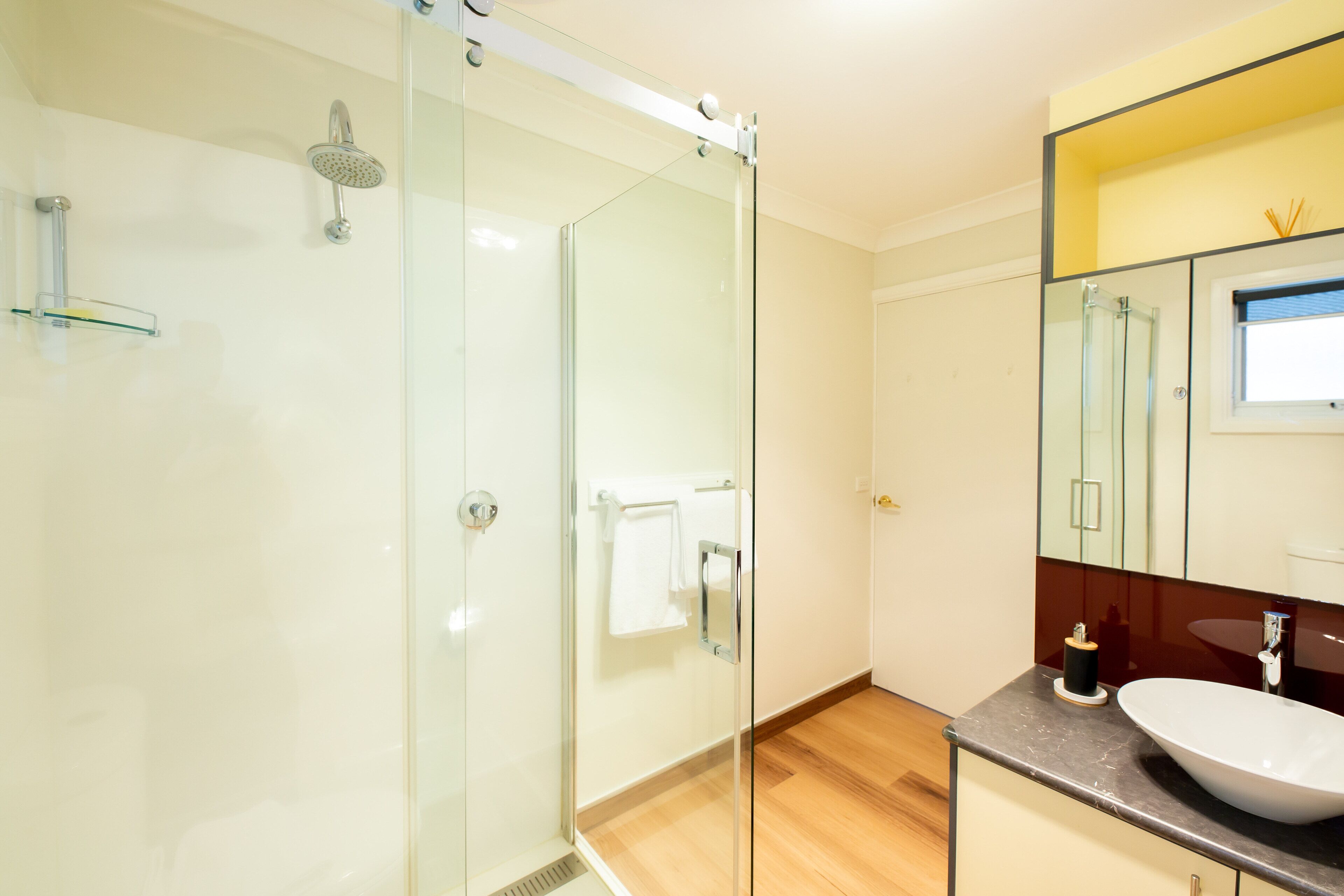 Ein modernes Badezimmer mit Duschkabine aus Glas, Holzboden, weißem Waschbecken und einem großen Spiegel.