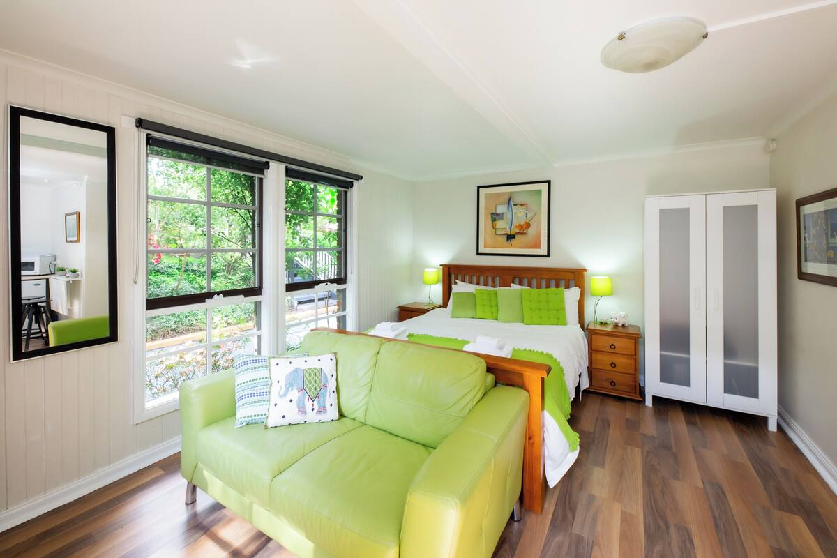 A bedroom with a bed, a green sofa, a nightstand, a wardrobe, and a window with a view of greenery.