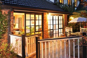 Eine Holzterrasse mit Geländer, ein Backsteinhaus mit großen Fenstern und eine Terrassenfläche mit Stühlen und Sonnenschirm.