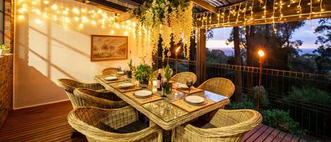 Un área de comedor al aire libre con sillas de mimbre, una mesa de vidrio, luces colgantes y vistas al exterior.