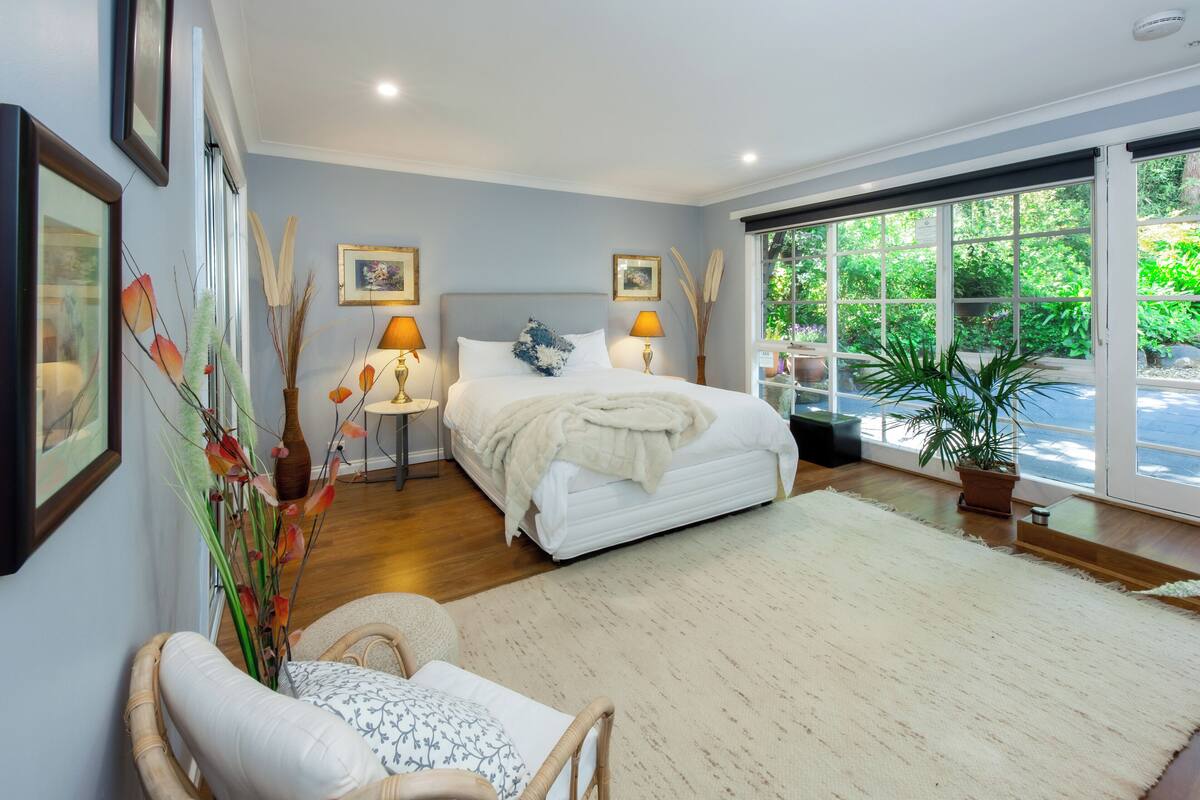 A bedroom with a bed, two bedside lamps, a chair, a large window, and a potted plant.