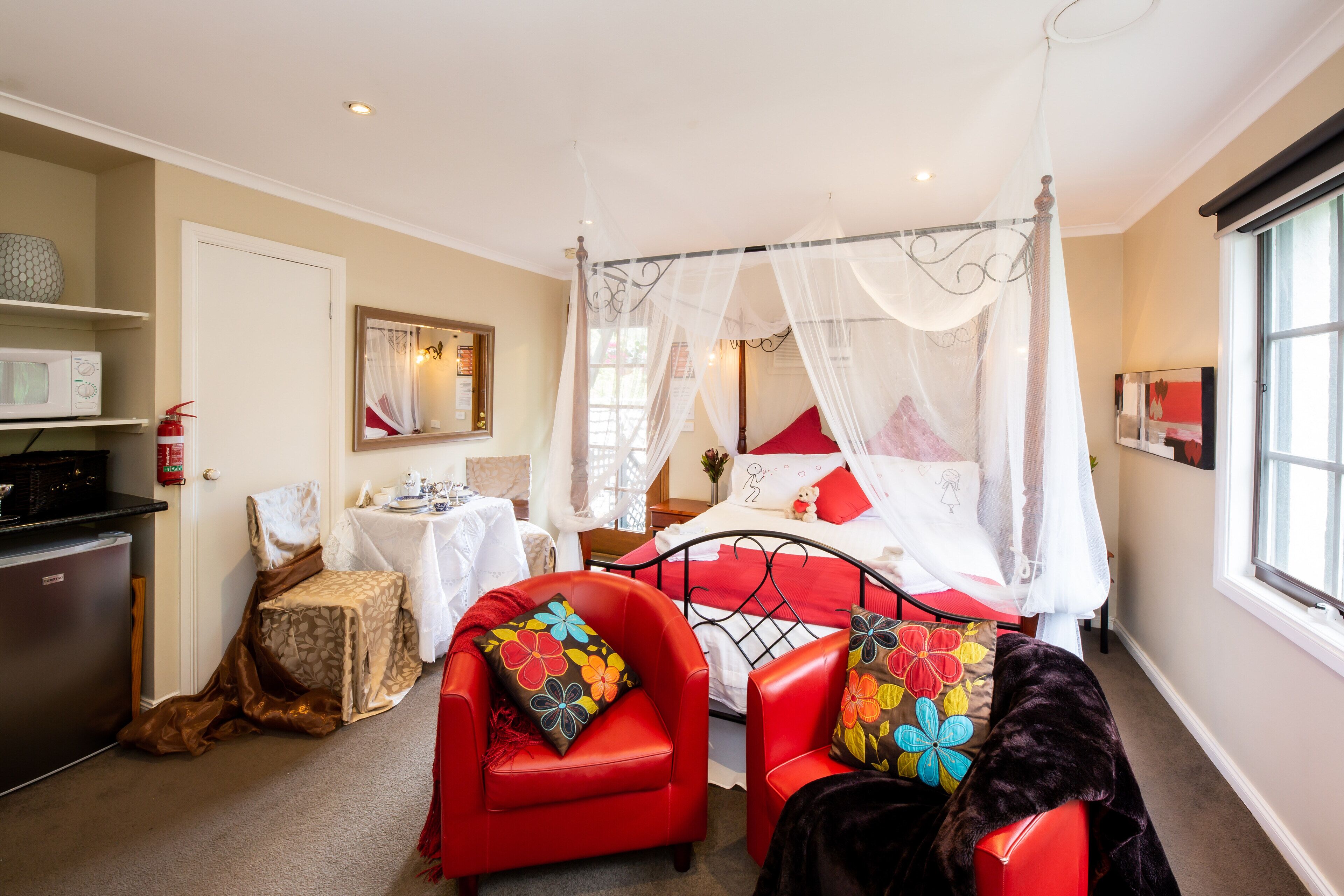 A four-poster canopy bed with a white canopy, red and white bedding, and colorful floral pillows. There are two red leather chairs with matching cushions. A small table with a white tablecloth and a vase of flowers is visible. A microwave and a fire extinguisher are on a counter to the left.