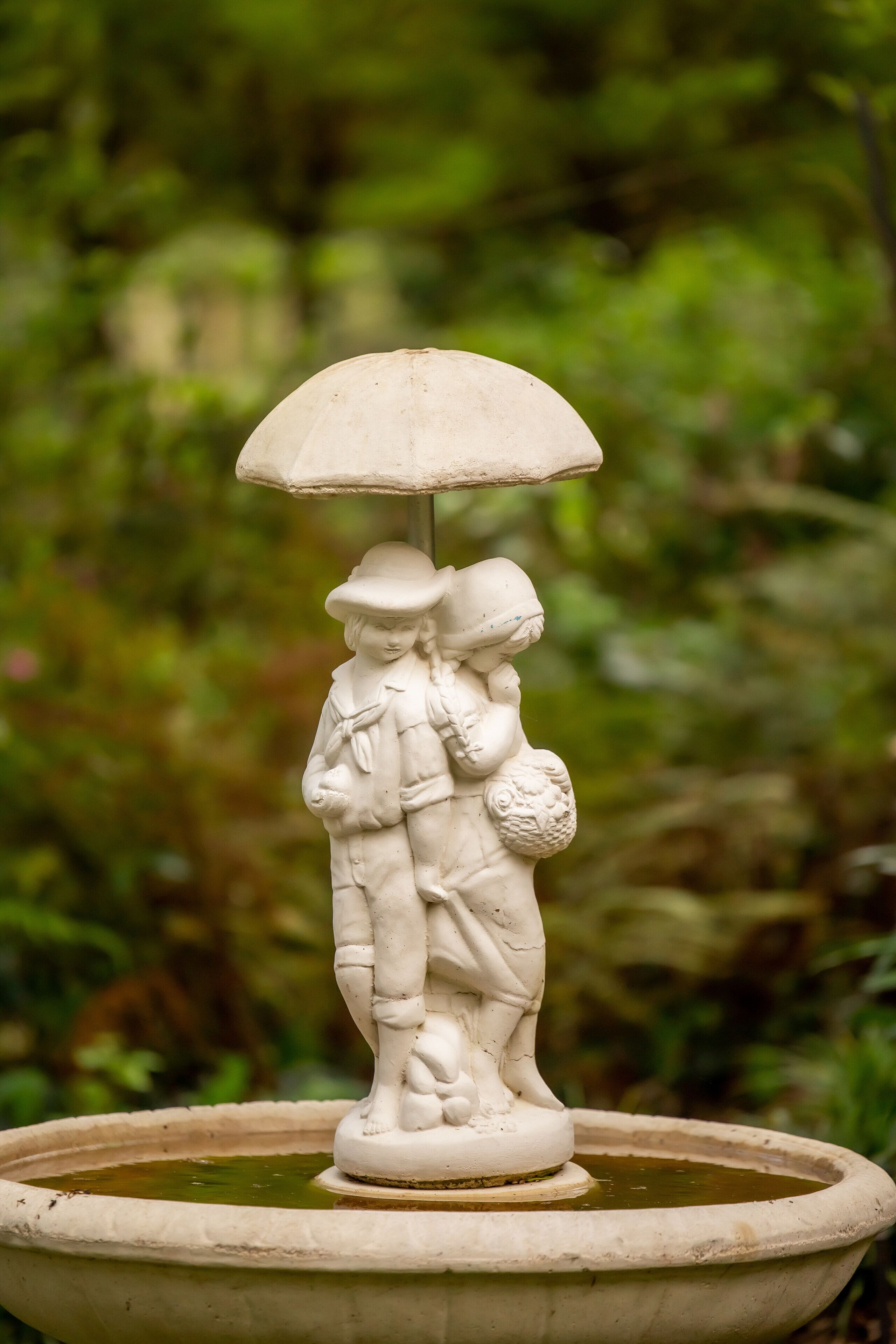 Une fontaine en pierre ornée d’une sculpture représentant un couple se tenant la main et tenant un parapluie, le tout entouré de verdure.