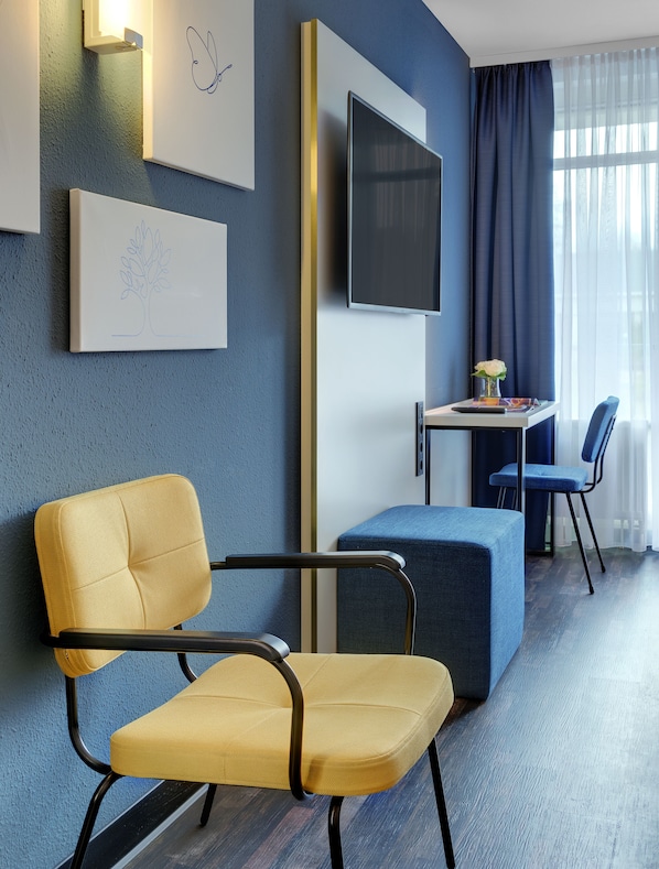 A modern hotel room with a blue textured wall, a flat-screen TV mounted above a desk, a yellow chair, a blue sofa, and a window with sheer curtains. - Hotel the YARD Bad Honnef (Bad Honnef)