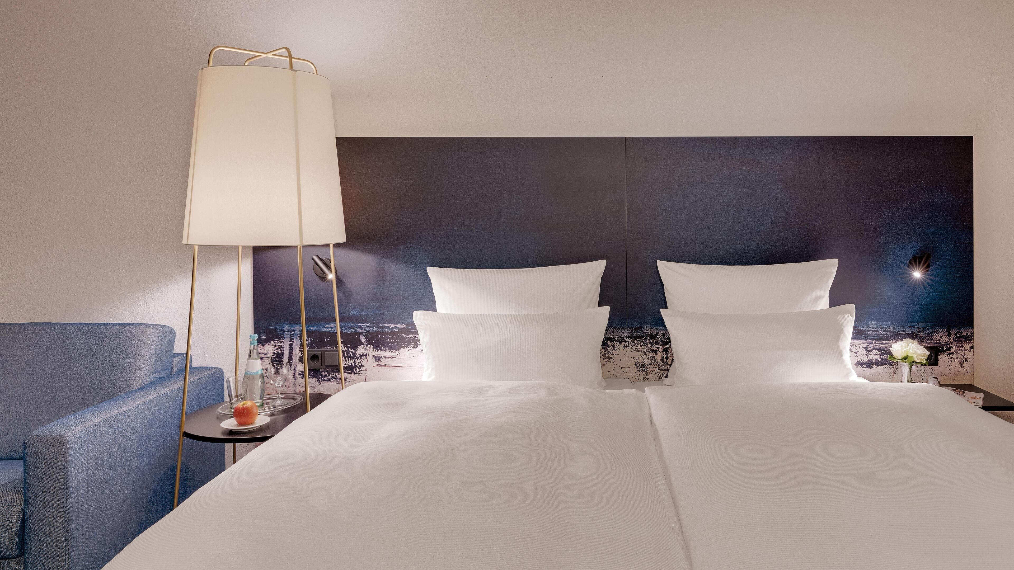 A hotel room with a large bed, a bedside table with a lamp, a small table with a bottle and glasses, and a blue armchair.