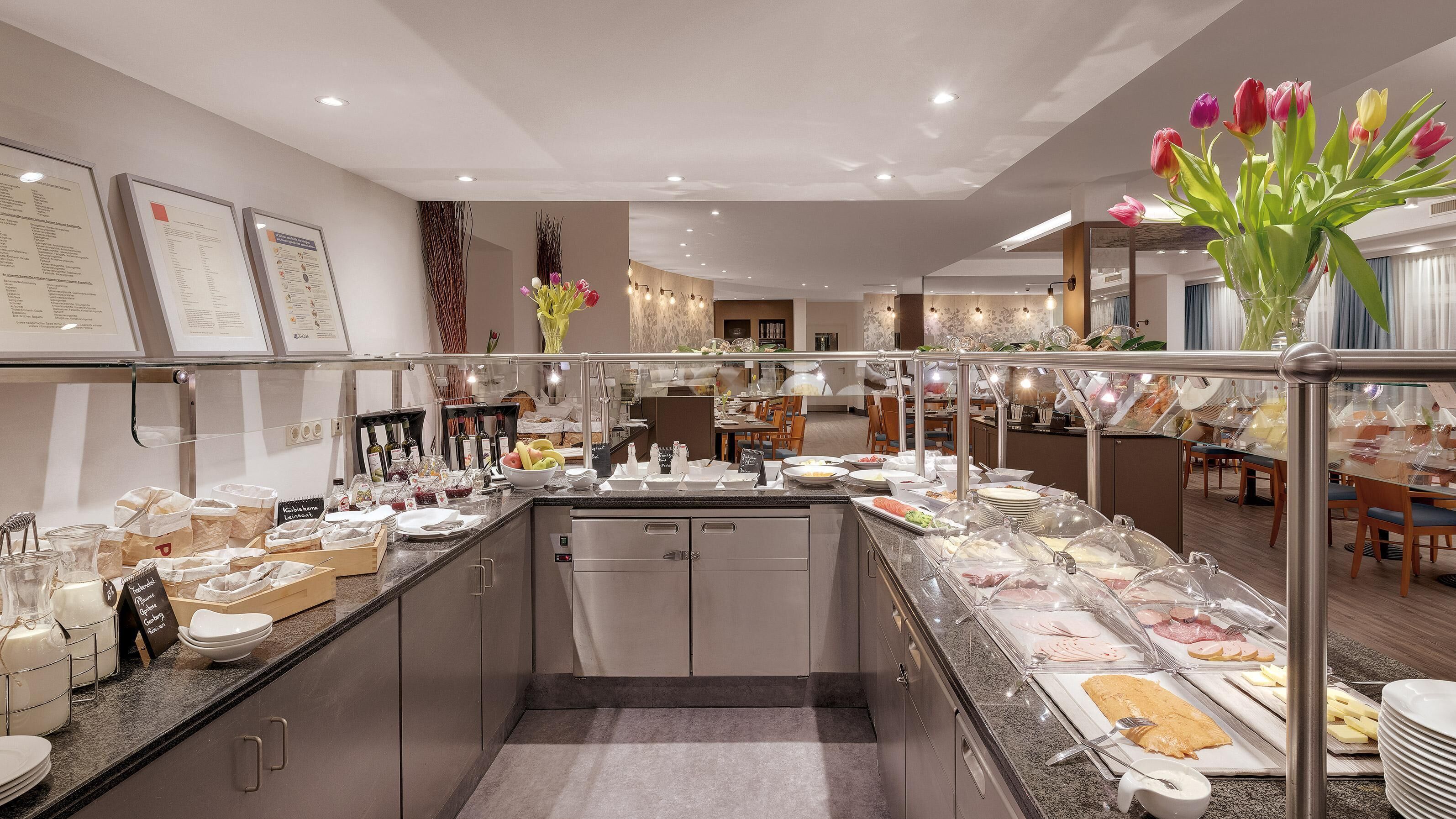 Un buffet de petit-déjeuner moderne, proposé dans un hôtel, avec un choix varié d’aliments, notamment des fruits, des fromages et des viandes, ainsi qu’un espace repas avec des tables et des chaises.