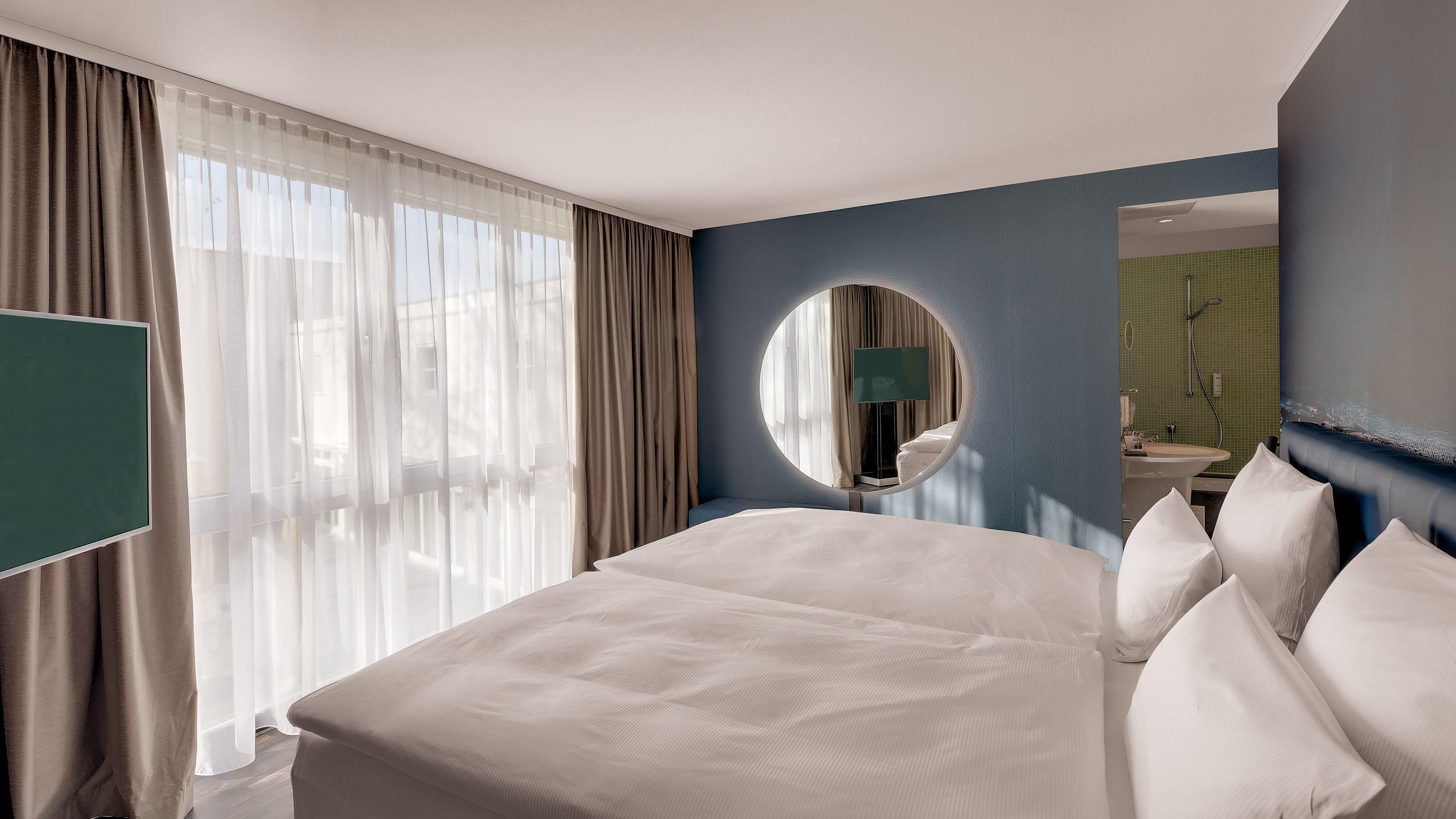 A hotel room with a large bed, a round mirror, a television, and a bathroom visible through an open door.