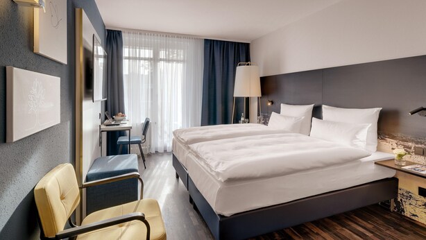 Habitación de hotel con una cama grande, un escritorio con una computadora portátil, una silla y vistas a la ciudad desde la ventana.