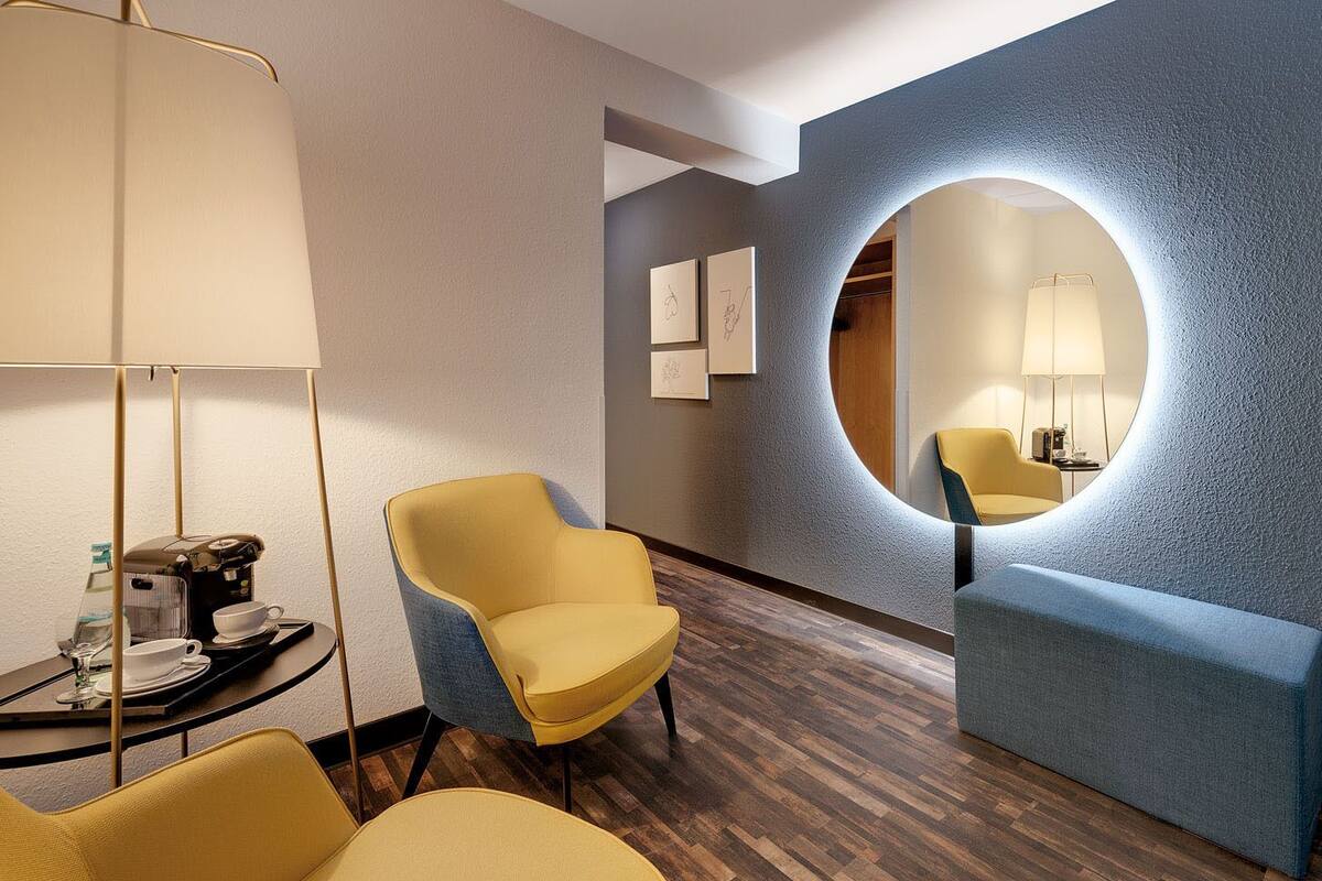 A modern hotel lobby with a round mirror, a yellow armchair, a blue bench, and a floor lamp.