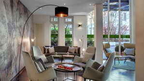 Una sala de estar moderna con un sofá, sillones, una mesa de centro con frutas y ventanas amplias con vistas a árboles y un edificio.