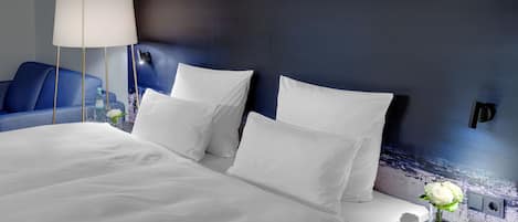 Una cama bien hecha con sábanas blancas, un sofá azul y una lámpara fijada a la pared.