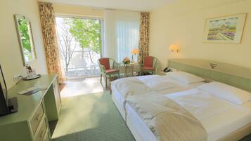 A hotel room with a large bed, a desk with a telephone, a chair, a small table with a lamp, and a window with curtains.