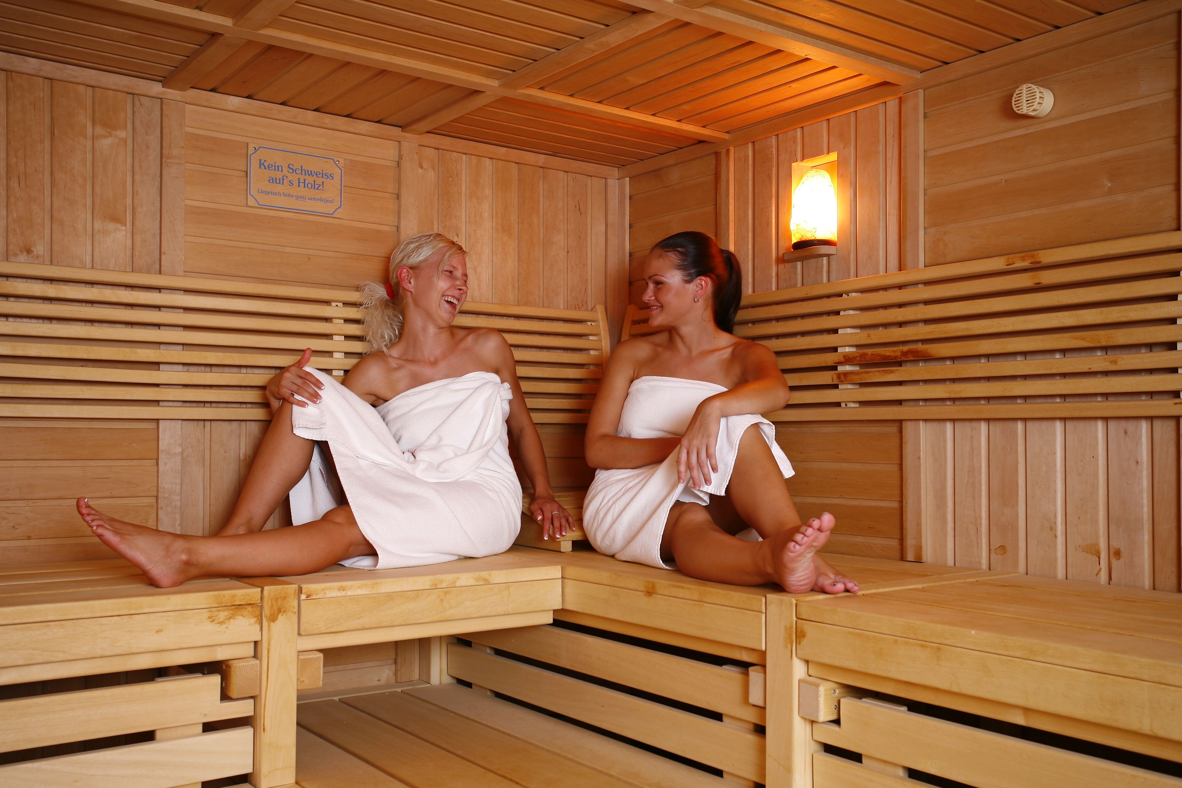Two people sitting in a wooden sauna with benches and a wall-mounted light.