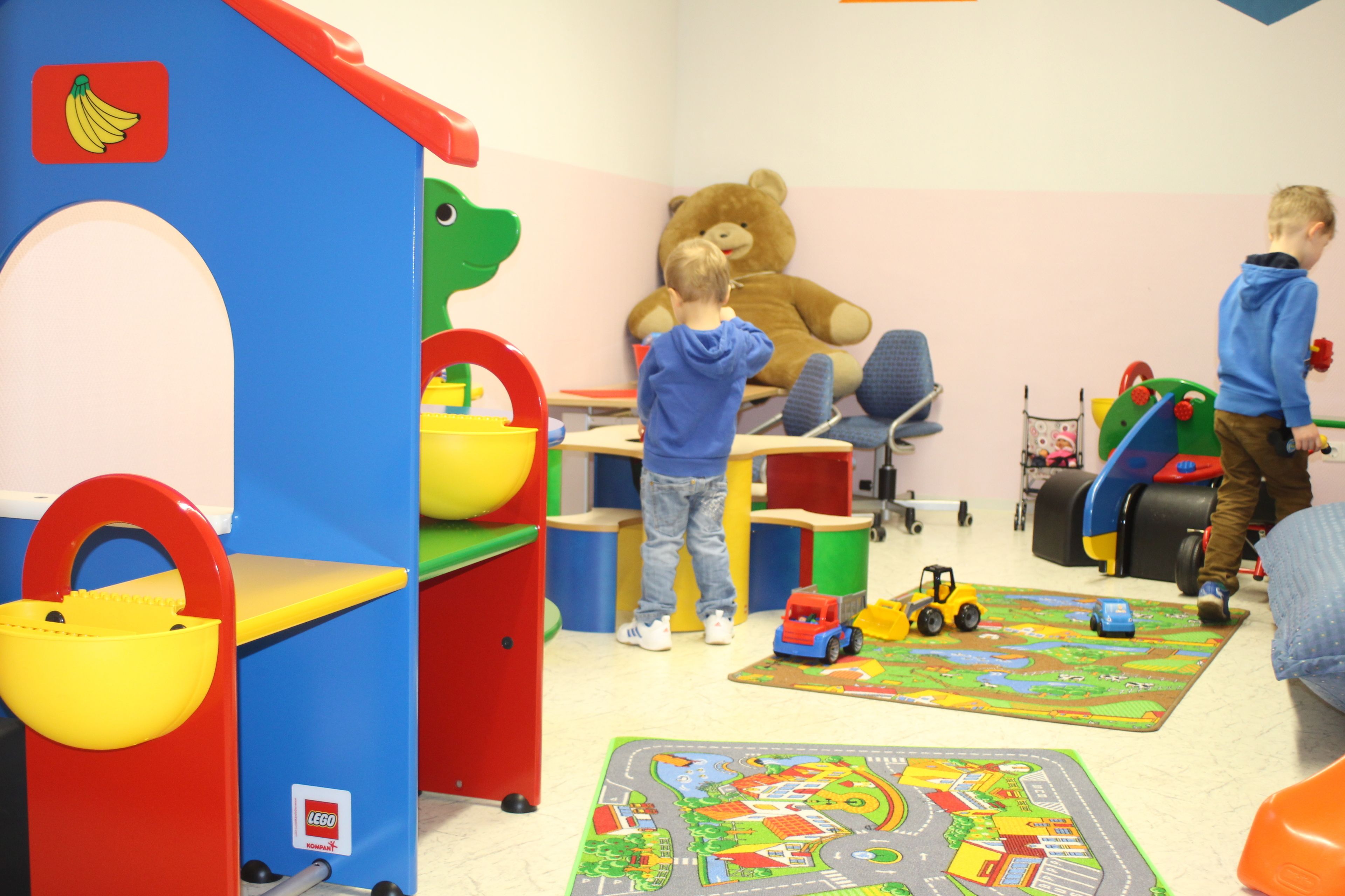 Un área de juegos para niños con un gran oso de peluche, una cocina de juguete y diversos juguetes.