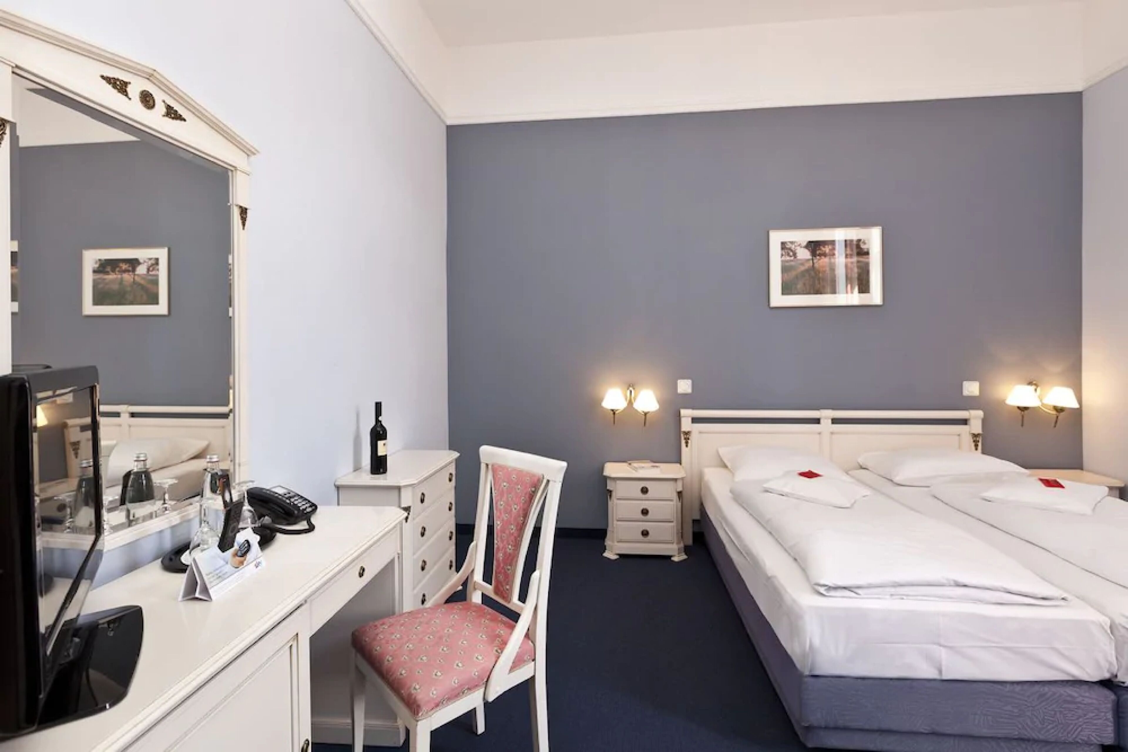 A hotel room with a desk, chair, two bedside tables, two beds, a mirror, and wall-mounted lights.