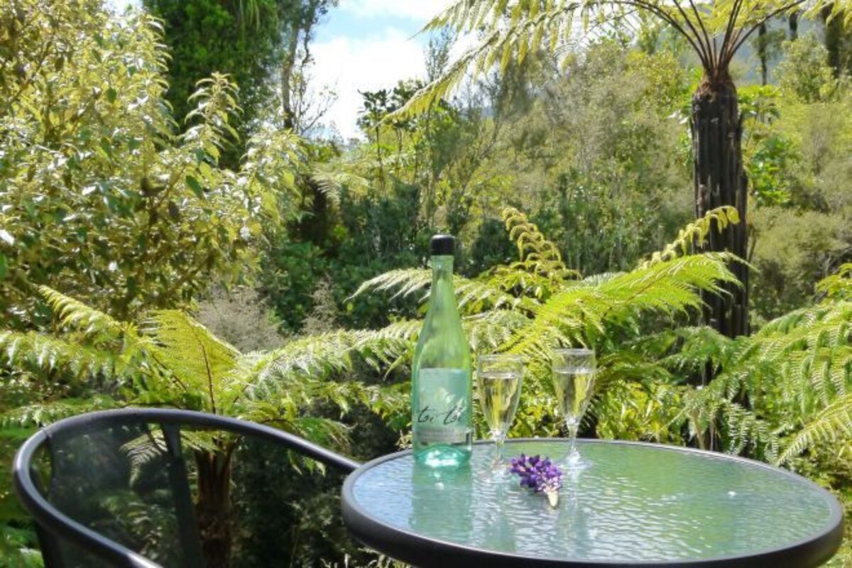 A glass table with a bottle and glasses, surrounded by lush greenery.