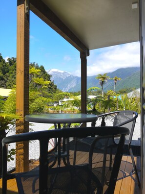 A covered outdoor seating area with a table and chairs, overlooking a mountainous landscape with greenery and a view of a snow-capped peak. - 10 Cottages (Franz Josef Glacier)