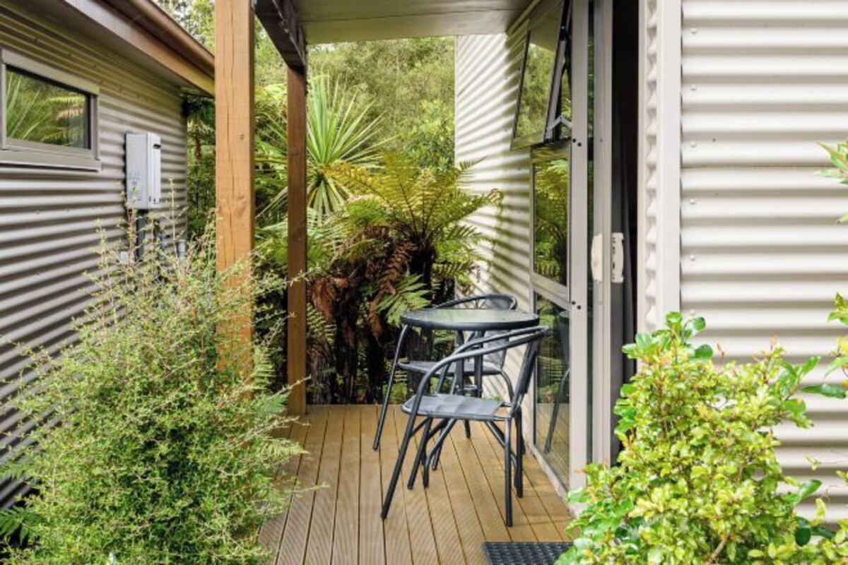 A wooden deck with a table and chairs, surrounded by lush greenery and a corrugated metal wall.