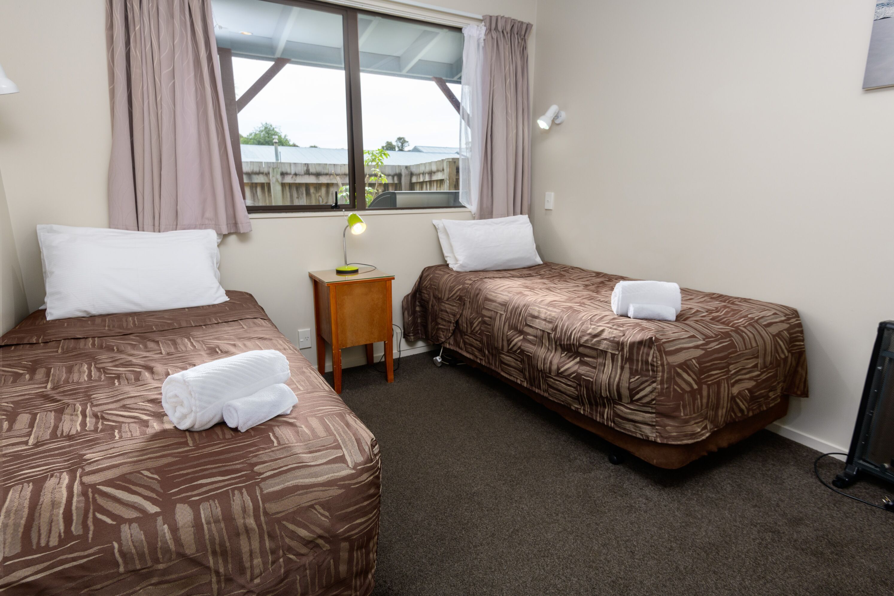 A hotel room with two beds, a small table with a lamp, and a window with curtains.