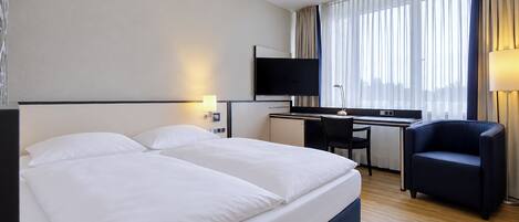 Quarto de hotel com uma cama grande, escrivaninha, cadeira, televisão e janela com cortinas.