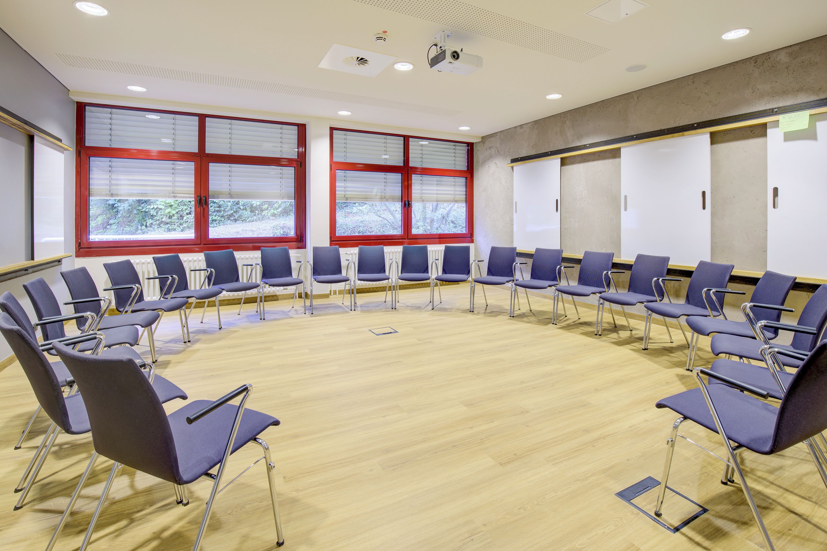 Ein Konferenzraum mit runder Sitzordnung, großen Fenstern mit Jalousien und einer Projektionsleinwand.