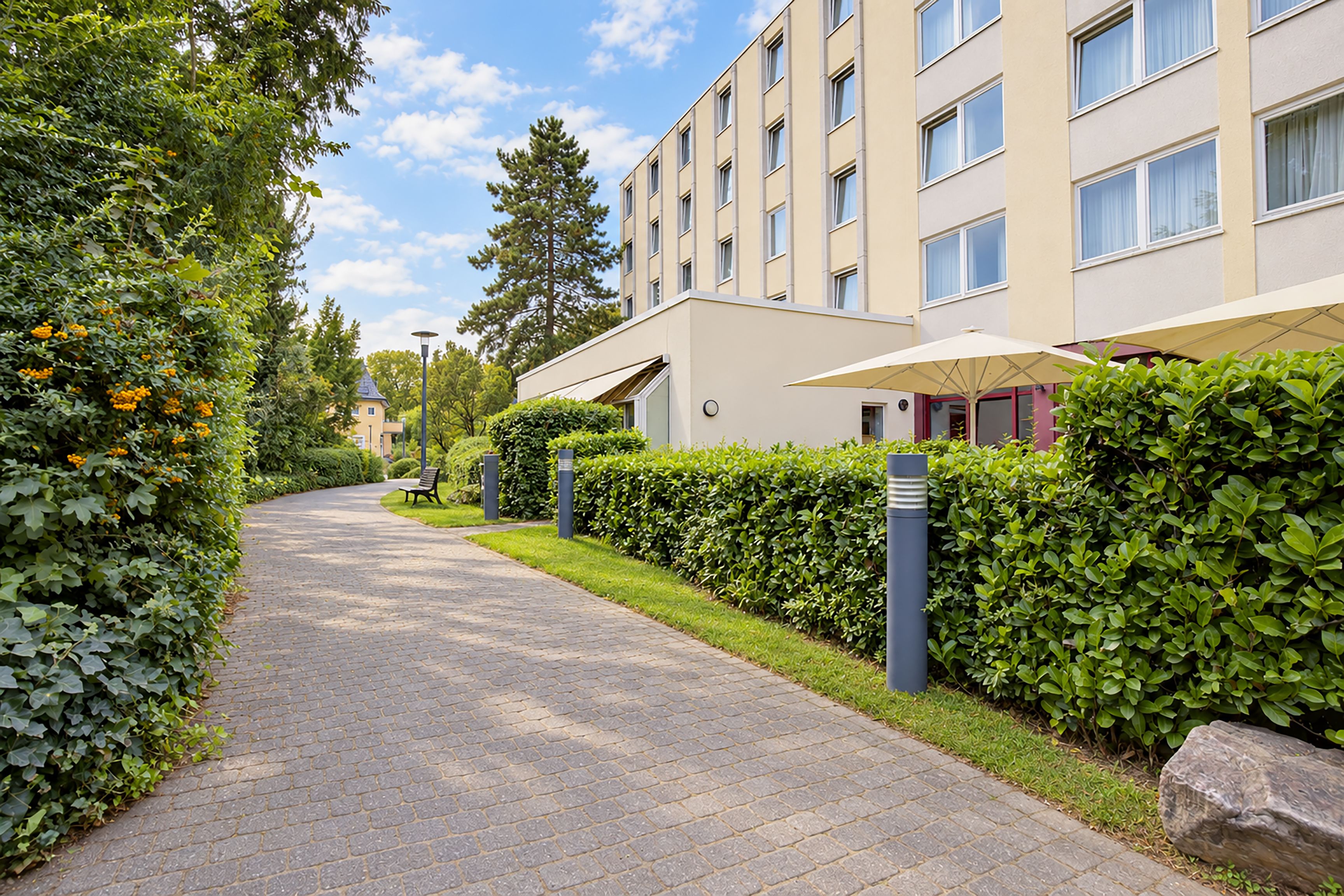 A pathway leading to a modern building with a flat roof, large windows, and a covered outdoor seating area.