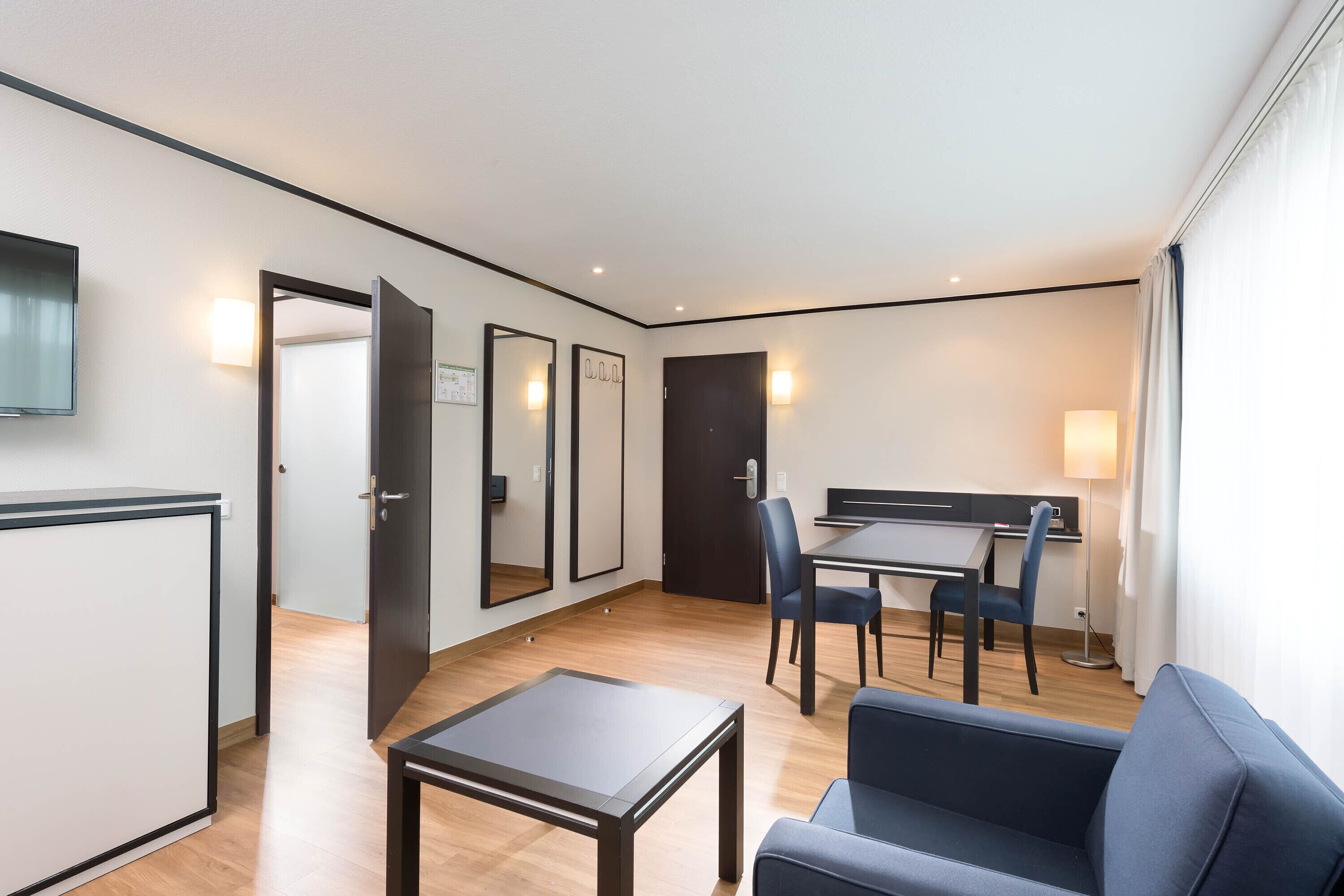 Una habitación de hotel moderna con una televisión de pantalla plana, un área de comedor con mesa y sillas, un sofá y un escritorio con lámpara.