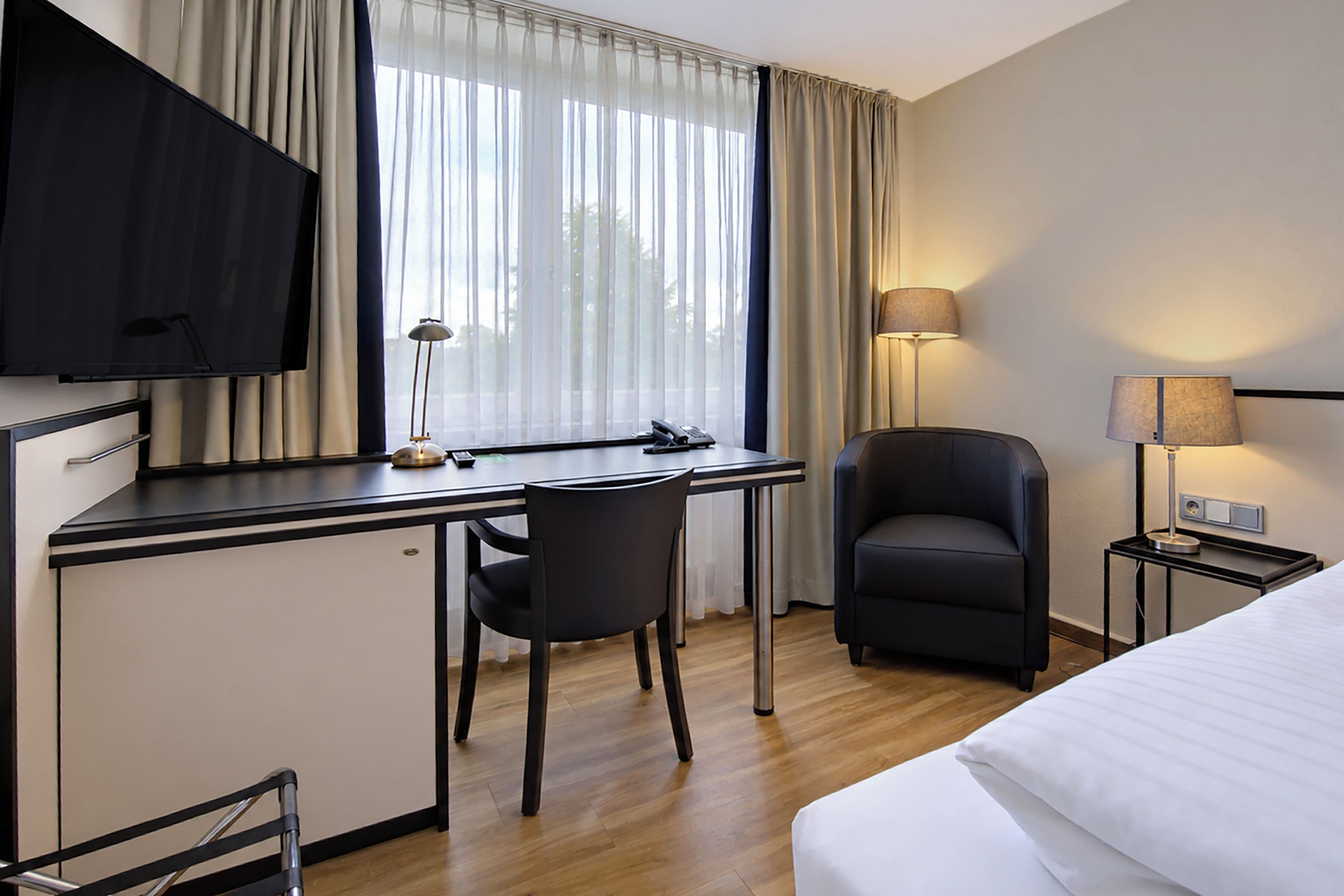 Ein Hotelzimmer mit einem Bett, einem Schreibtisch mit Stuhl, einem Fernseher, einer Lampe und einem Fenster mit Vorhängen.