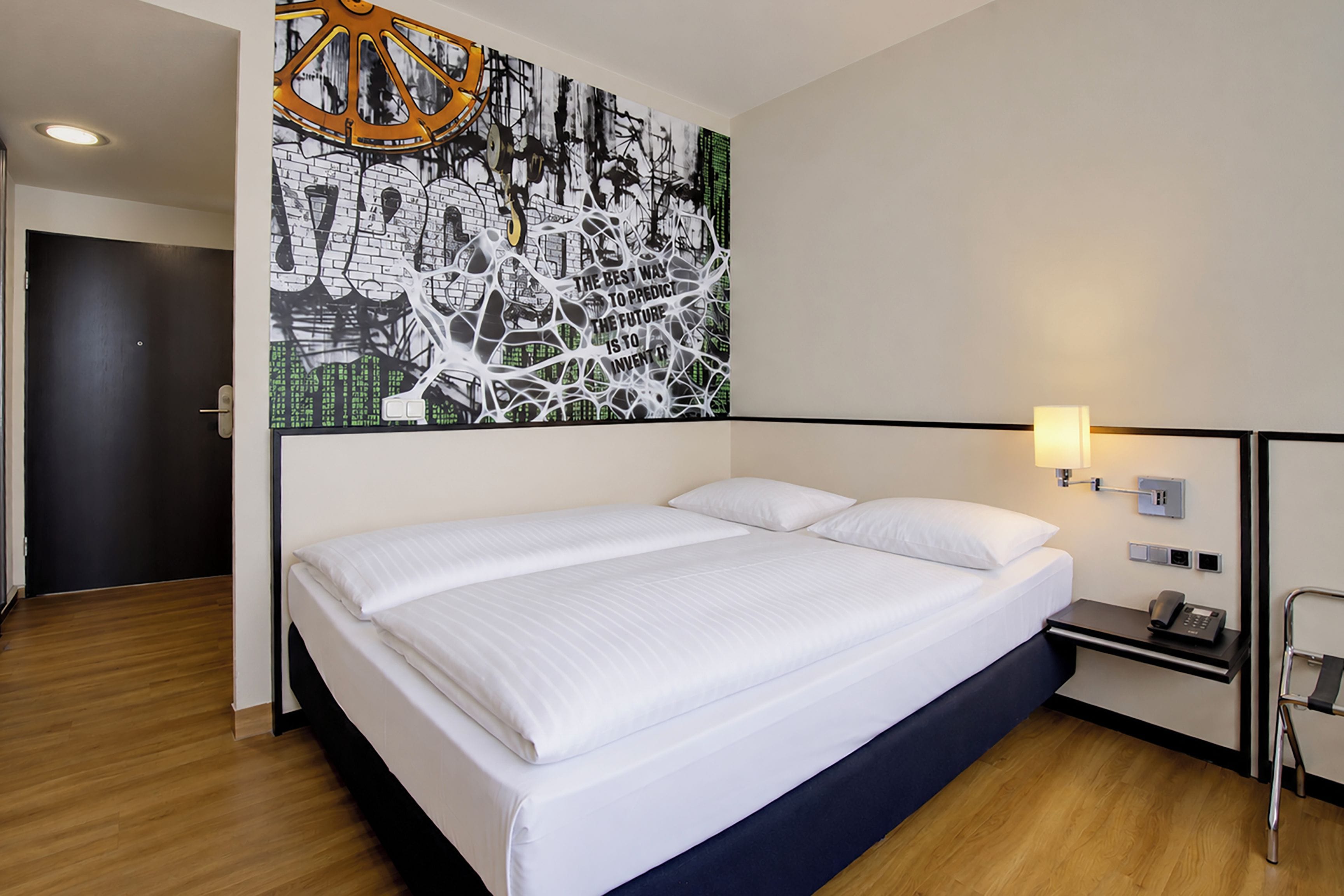 Une chambre d’hôtel avec un grand lit, une lampe fixée au mur et une œuvre d’art contemporaine.