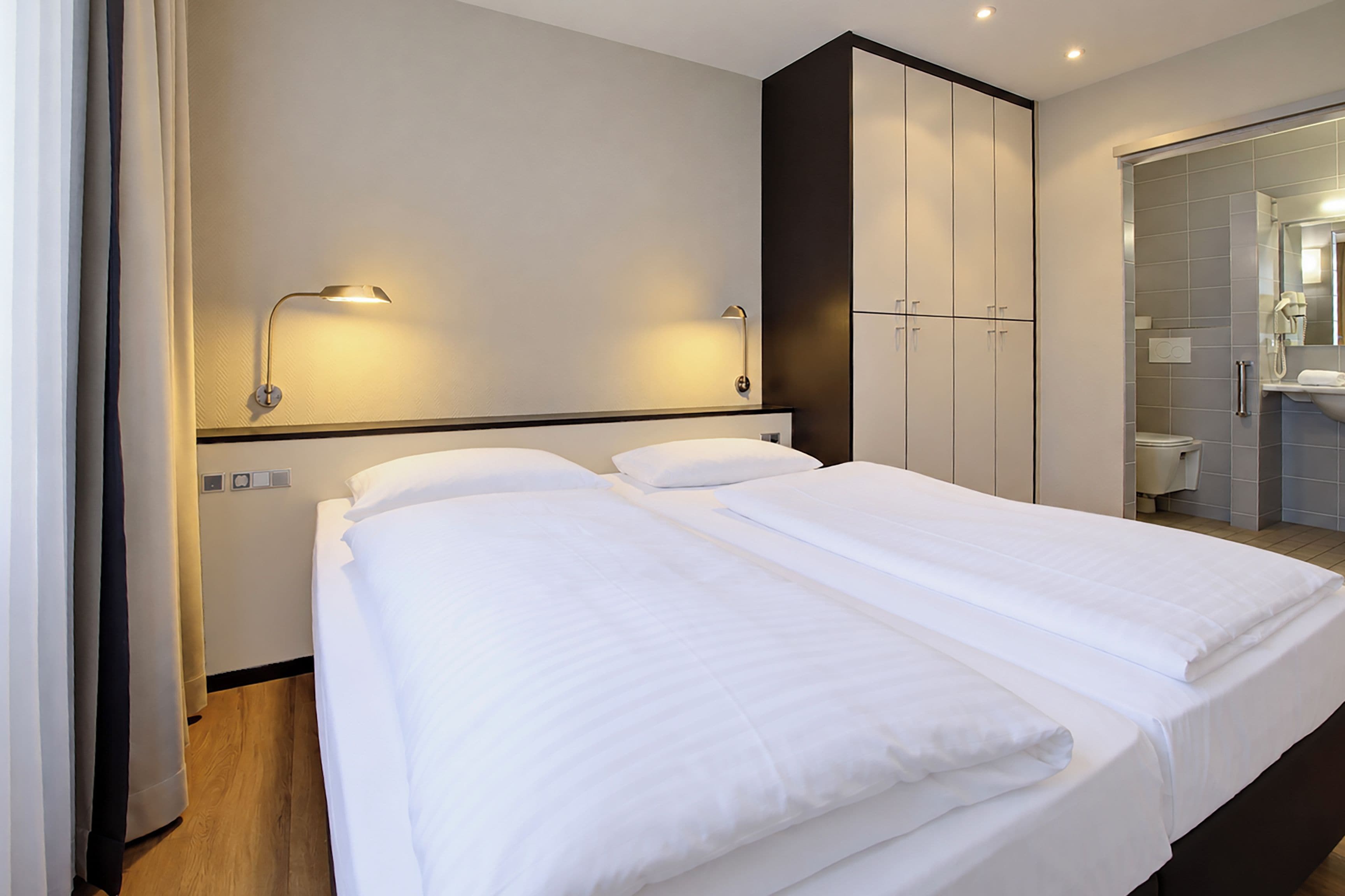 Habitación de hotel con dos camas, un armario, un baño con espejo y dos lámparas de pared.