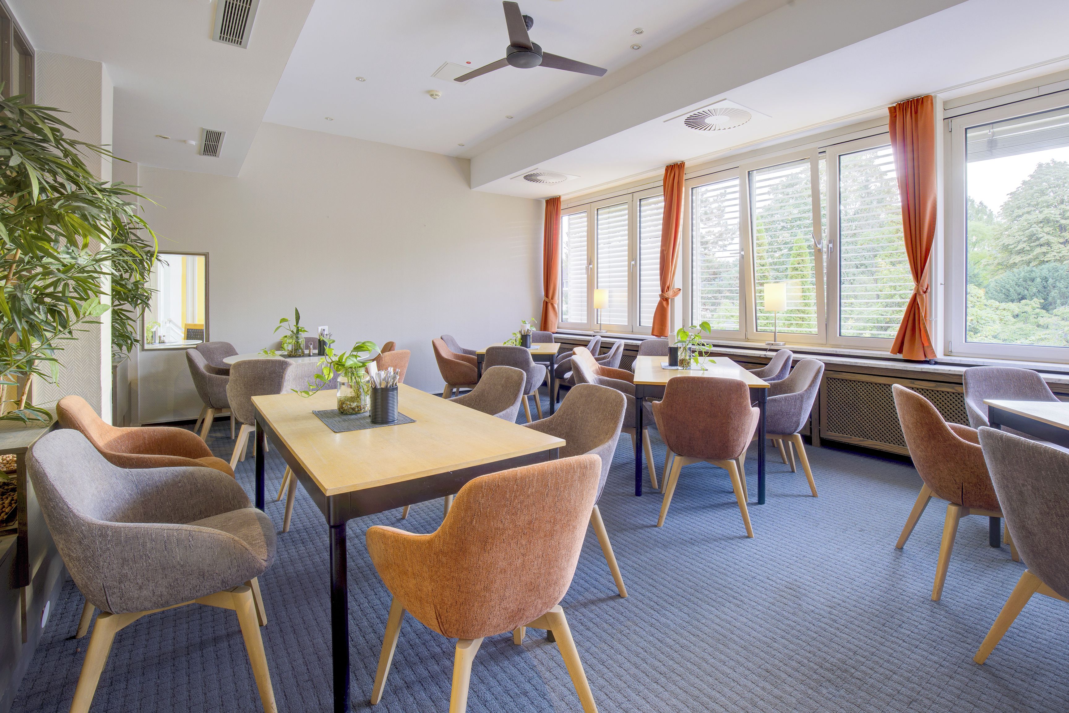 Een vergaderzaal met een lange tafel, meerdere stoelen, een plafondventilator en grote ramen met gordijnen.