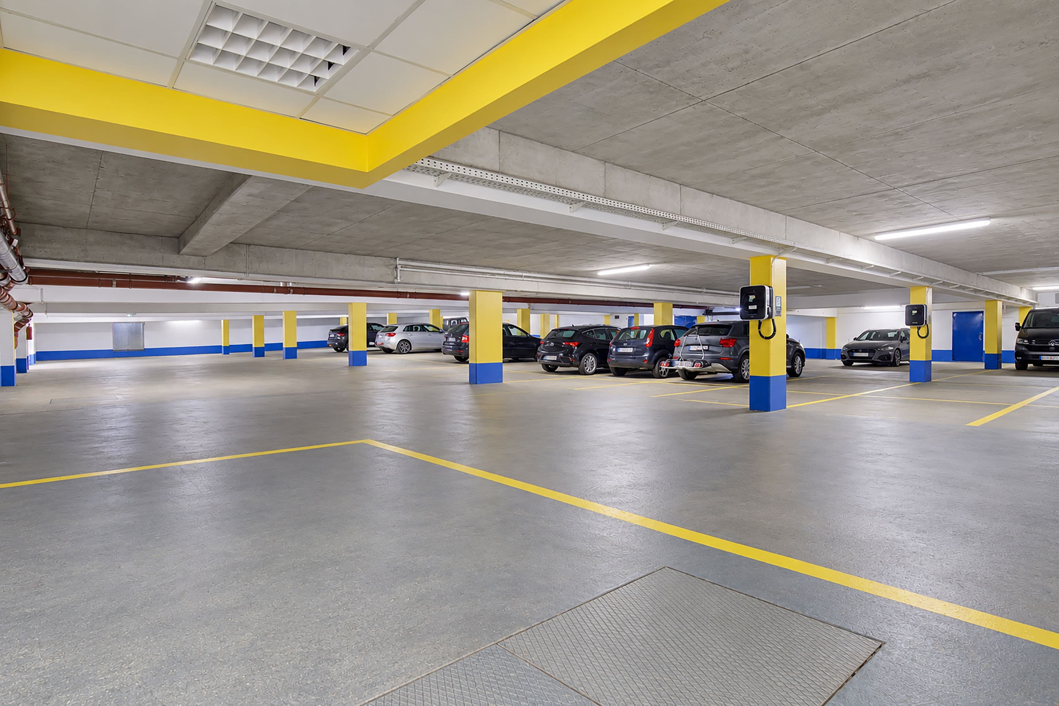 Un pàrquing subterrani de diversos nivells, amb pilars grocs i blaus, sòls de formigó i punts de càrrega per a vehicles elèctrics.