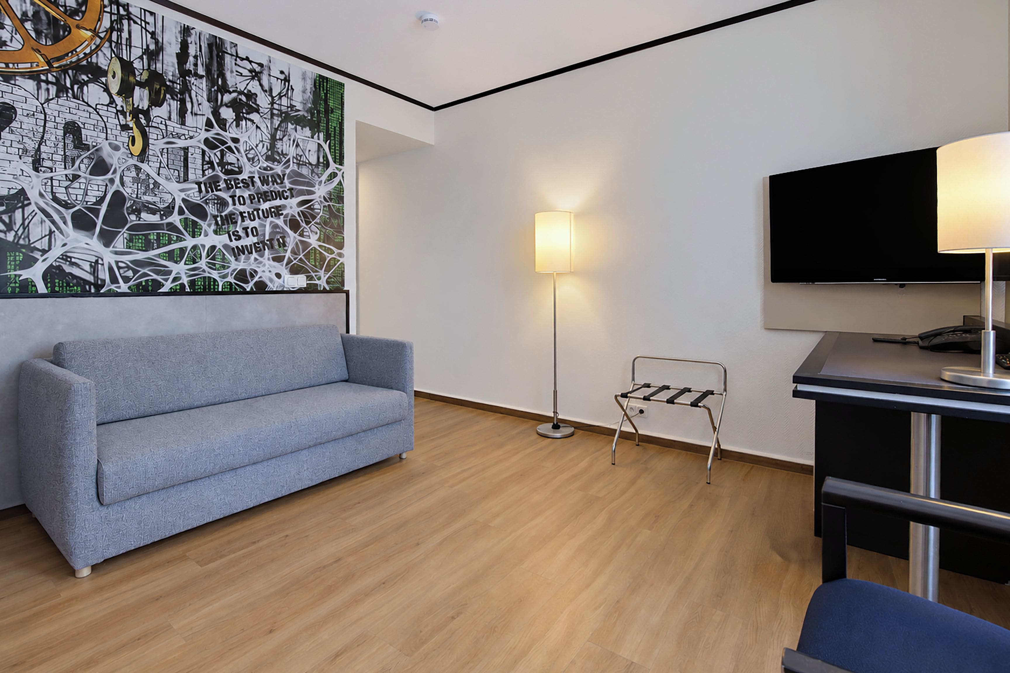 A modern hotel room with a grey sofa, a floor lamp, a TV, and a wall art piece.
