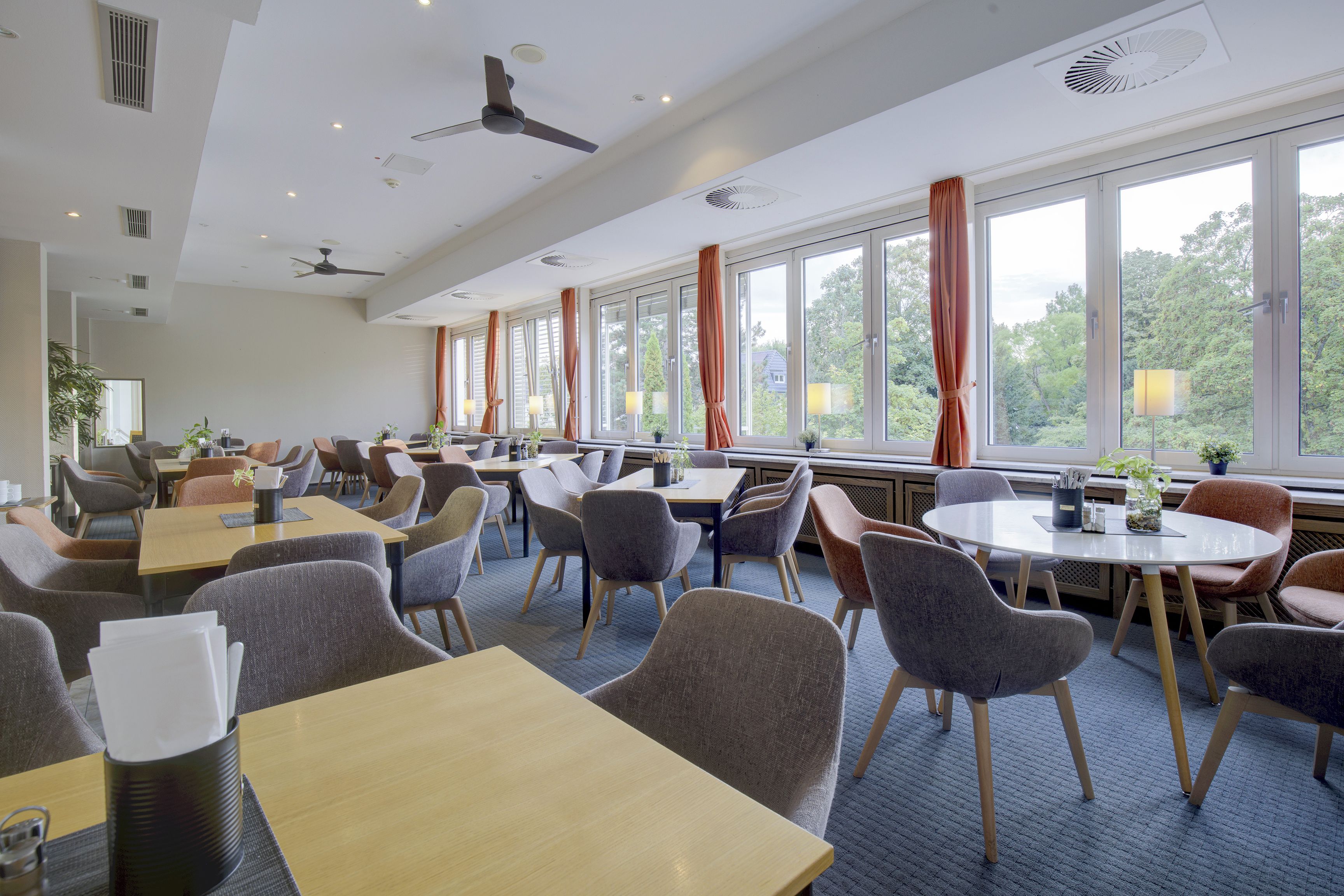 A hotel dining area with multiple tables and chairs, large windows with curtains, and ceiling fans.