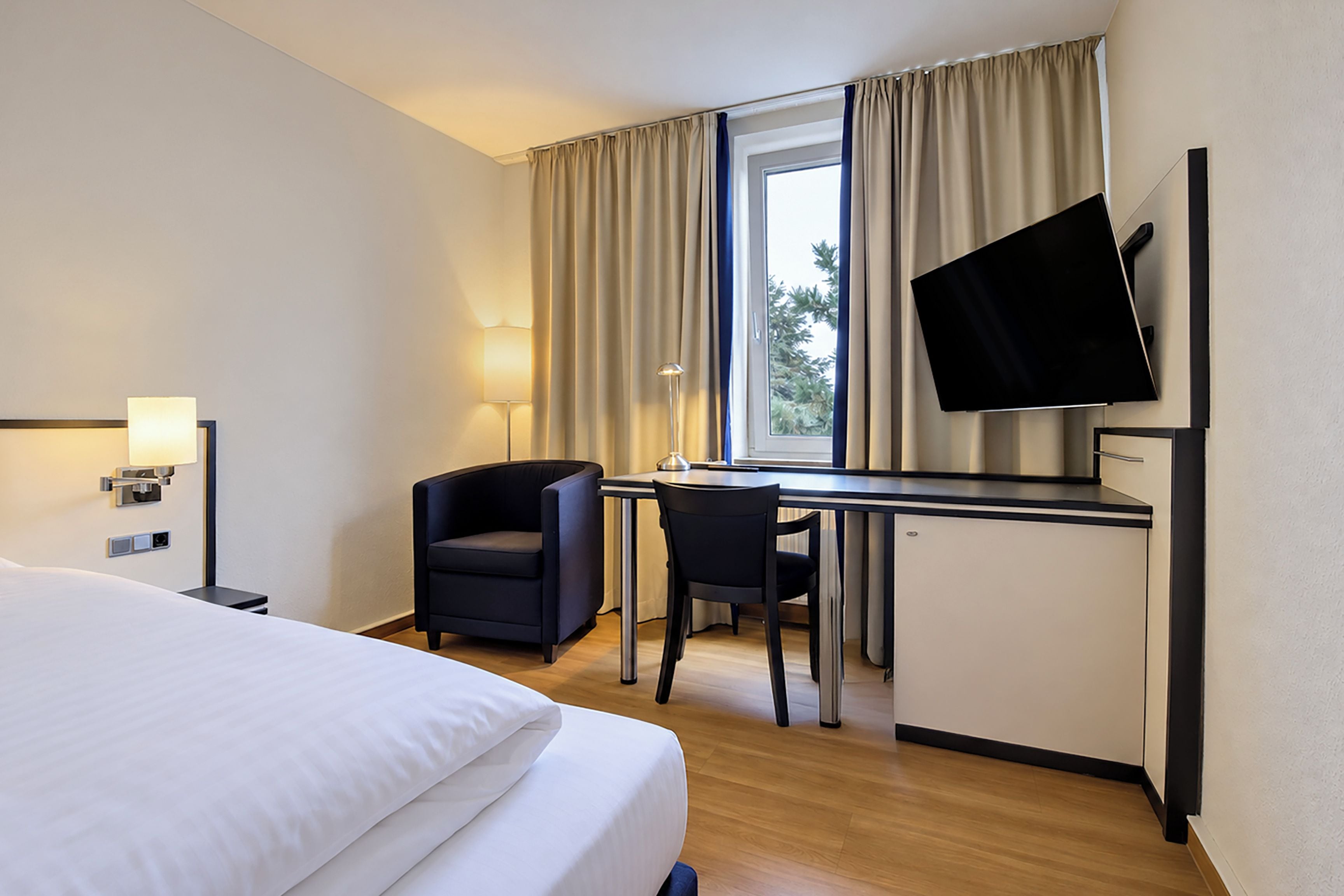 A hotel room with a bed, desk, chair, TV, and window with curtains.
