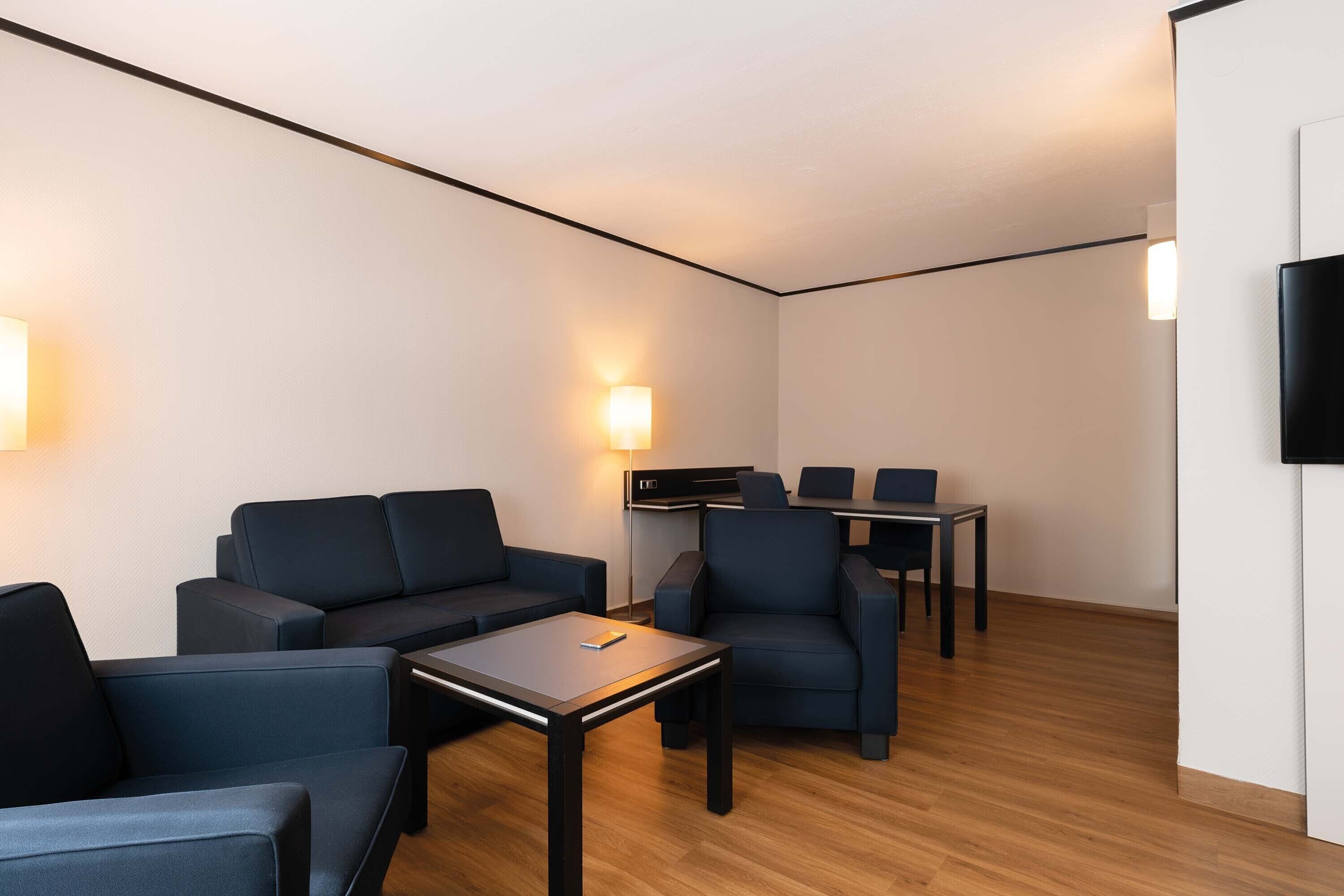 A modern, well-lit room with a sofa, chairs, a small table, and a TV mounted on the wall.