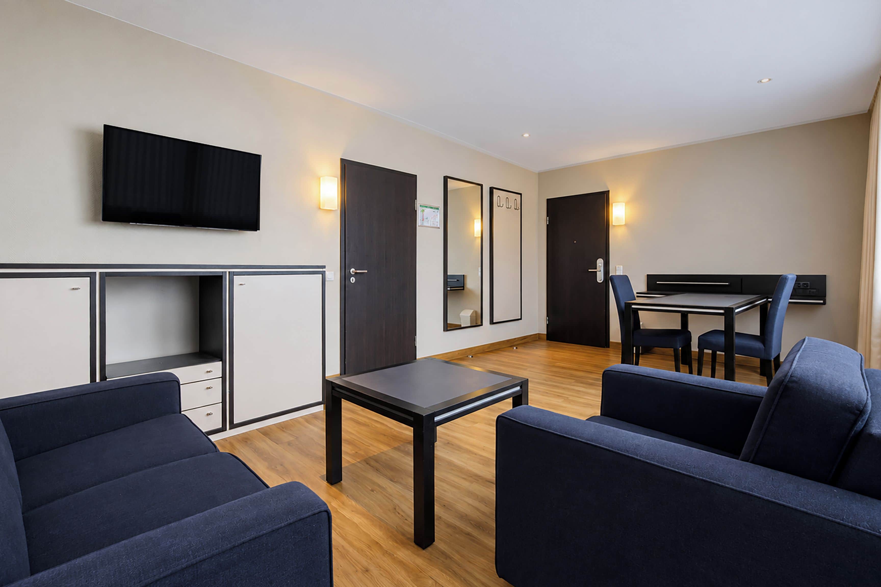 A modern hotel room with a flat-screen TV, a desk with a chair, a sofa, and a wardrobe.