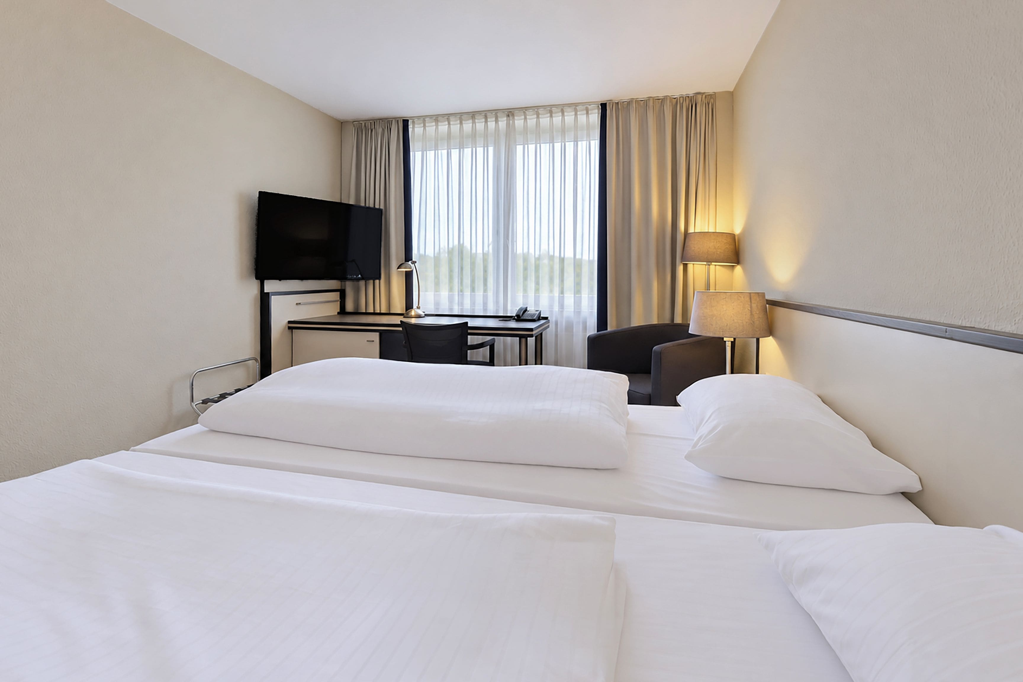 Ein Hotelzimmer mit einem Bett, einem Schreibtisch, einem Stuhl, einem Fernseher und einem Fenster mit Vorhängen.