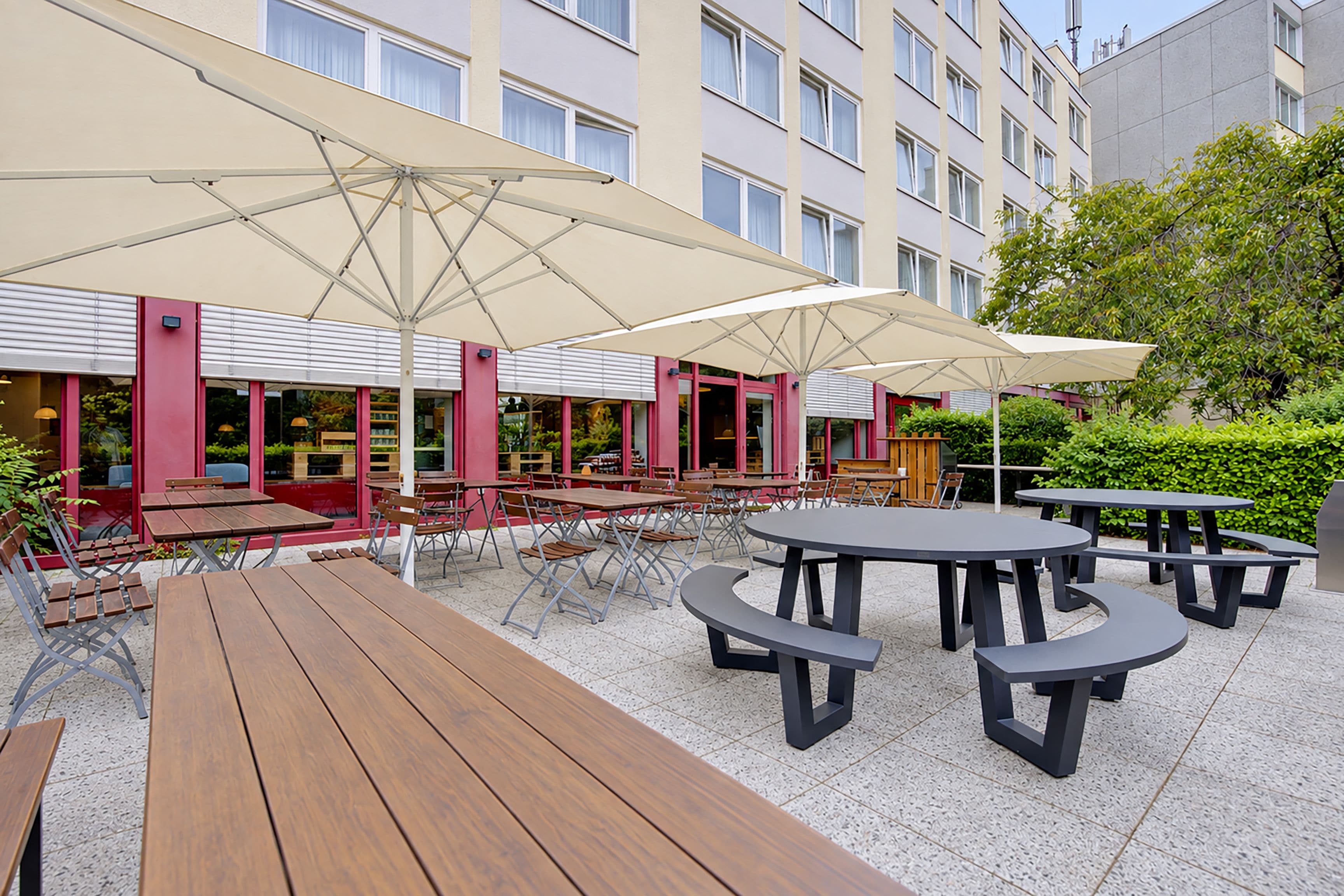 Ein Terrassenbereich im Freien mit mehreren Tischen und Stühlen, großen Sonnenschirmen und einem Gebäude mit zahlreichen Fenstern im Hintergrund.