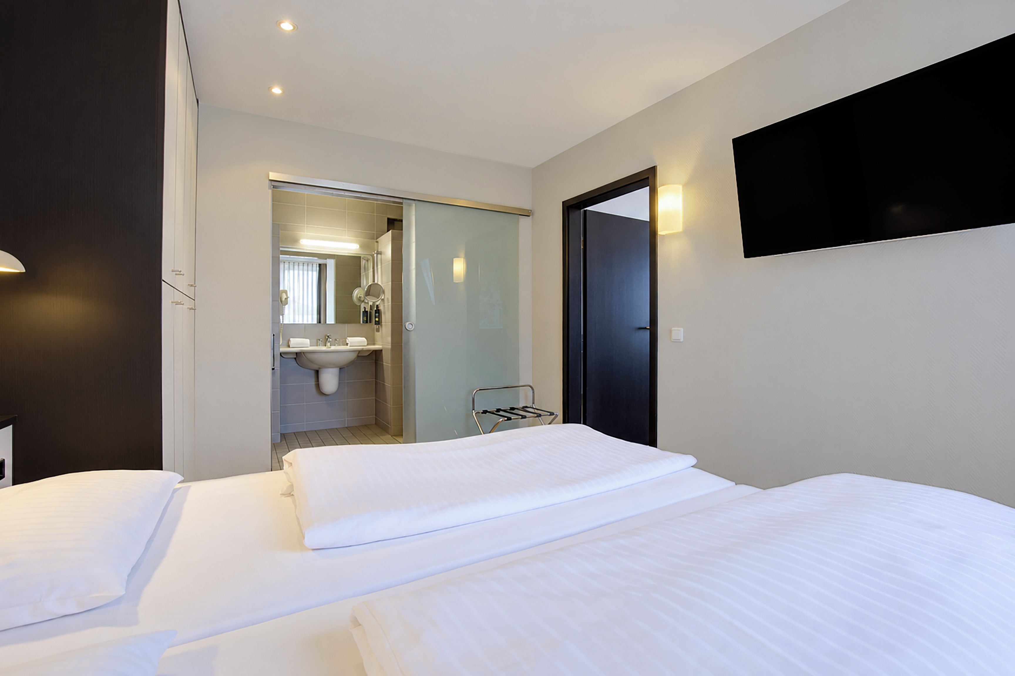 Ein Hotelzimmer mit einem Bett, einem Nachttisch, einem flachbildfernseher an der Wand und einem durch eine offene Tür einsehbaren Badezimmer.