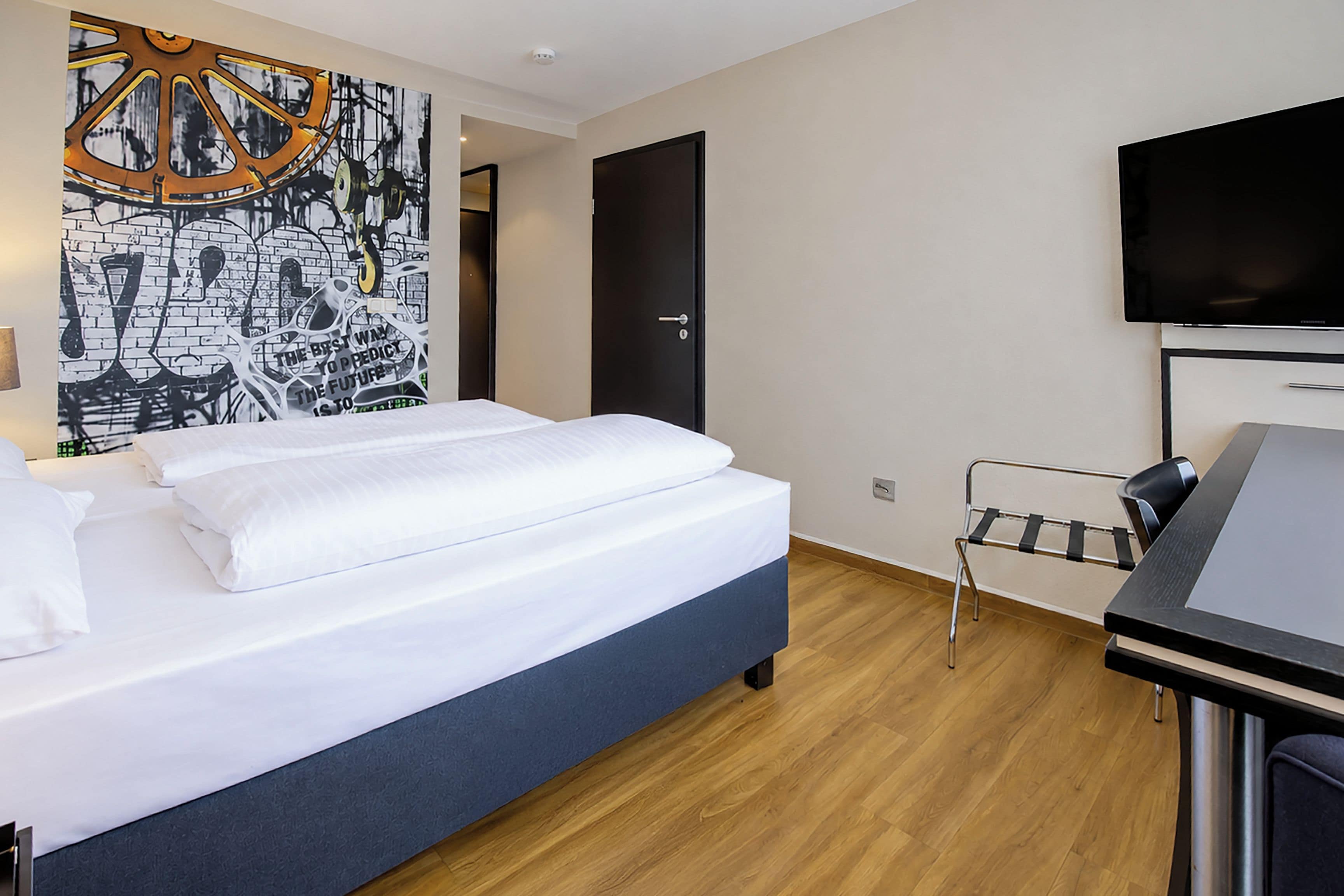 Une chambre d’hôtel équipée d’un lit, d’un bureau, d’une chaise, d’une télévision et d’un mur orné d’un motif graphique.