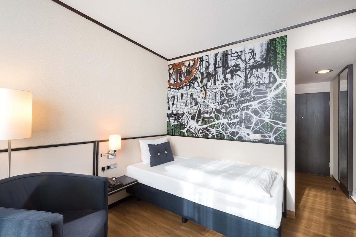 A hotel room with a bed, bedside table, lamp, and a large abstract painting on the wall.