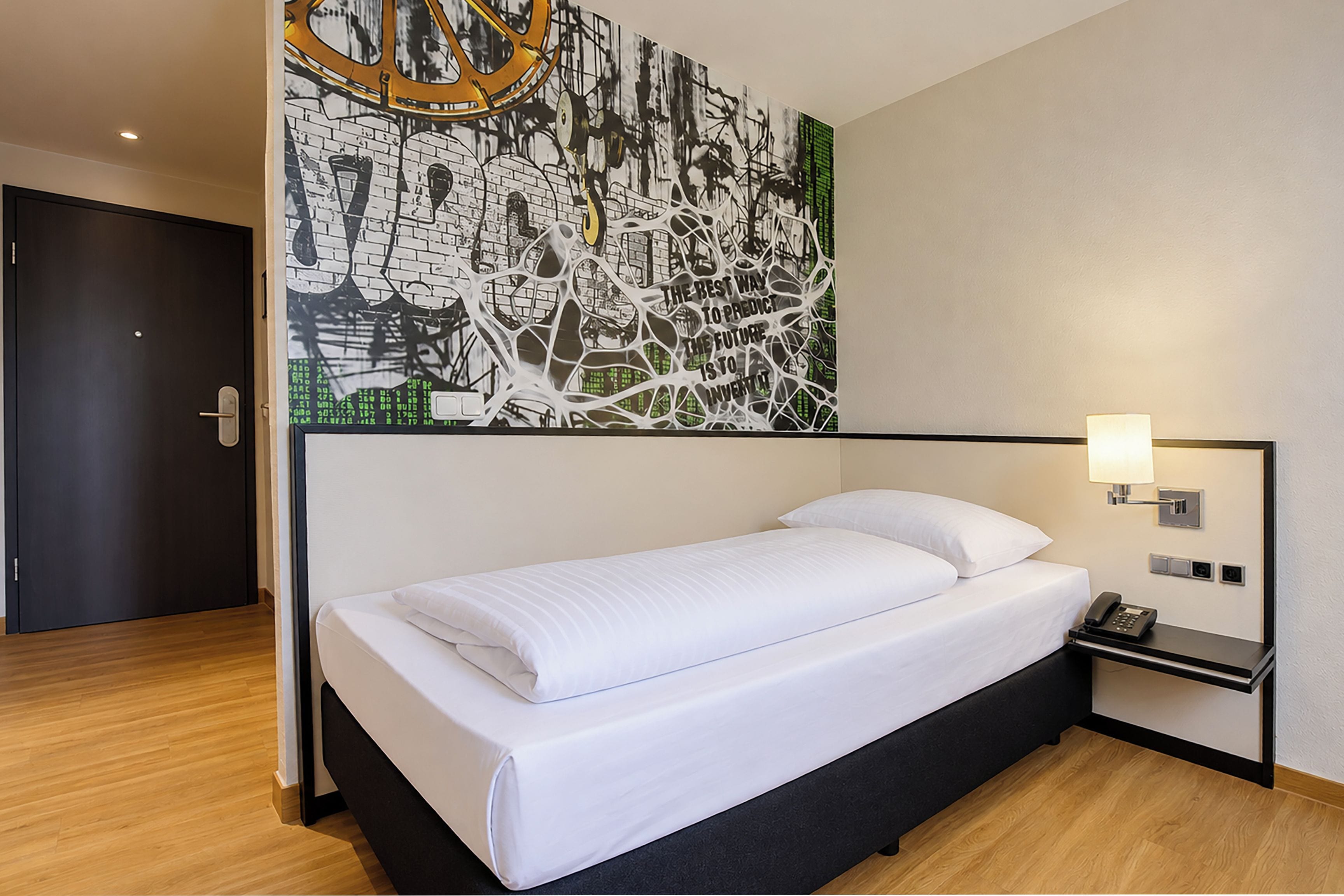 Een hotelkamer met een enkel bed, een nachtkastje met een lamp, en een groot abstract schilderij aan de muur.