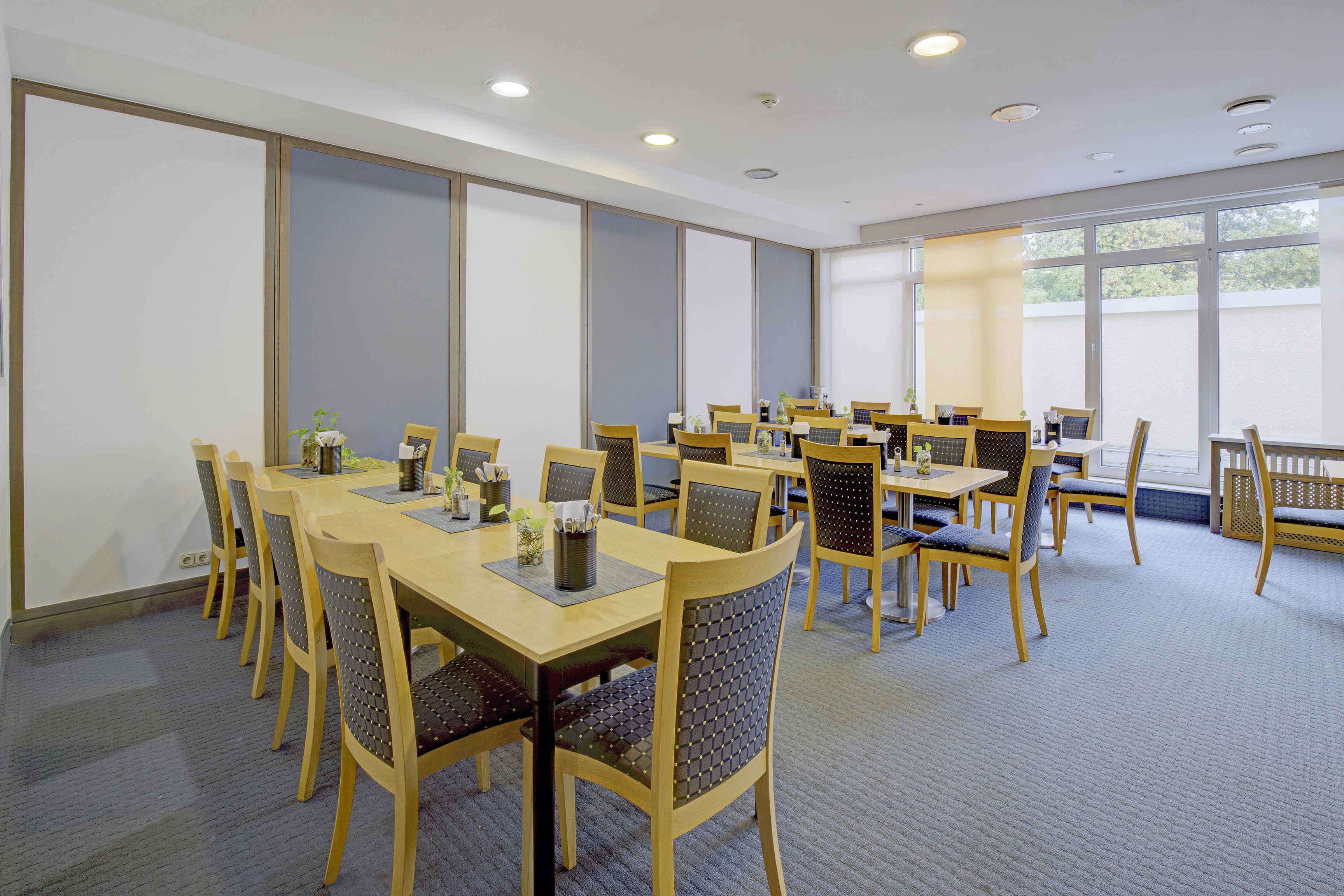 A conference room with multiple tables and chairs, large windows, and ceiling lights.
