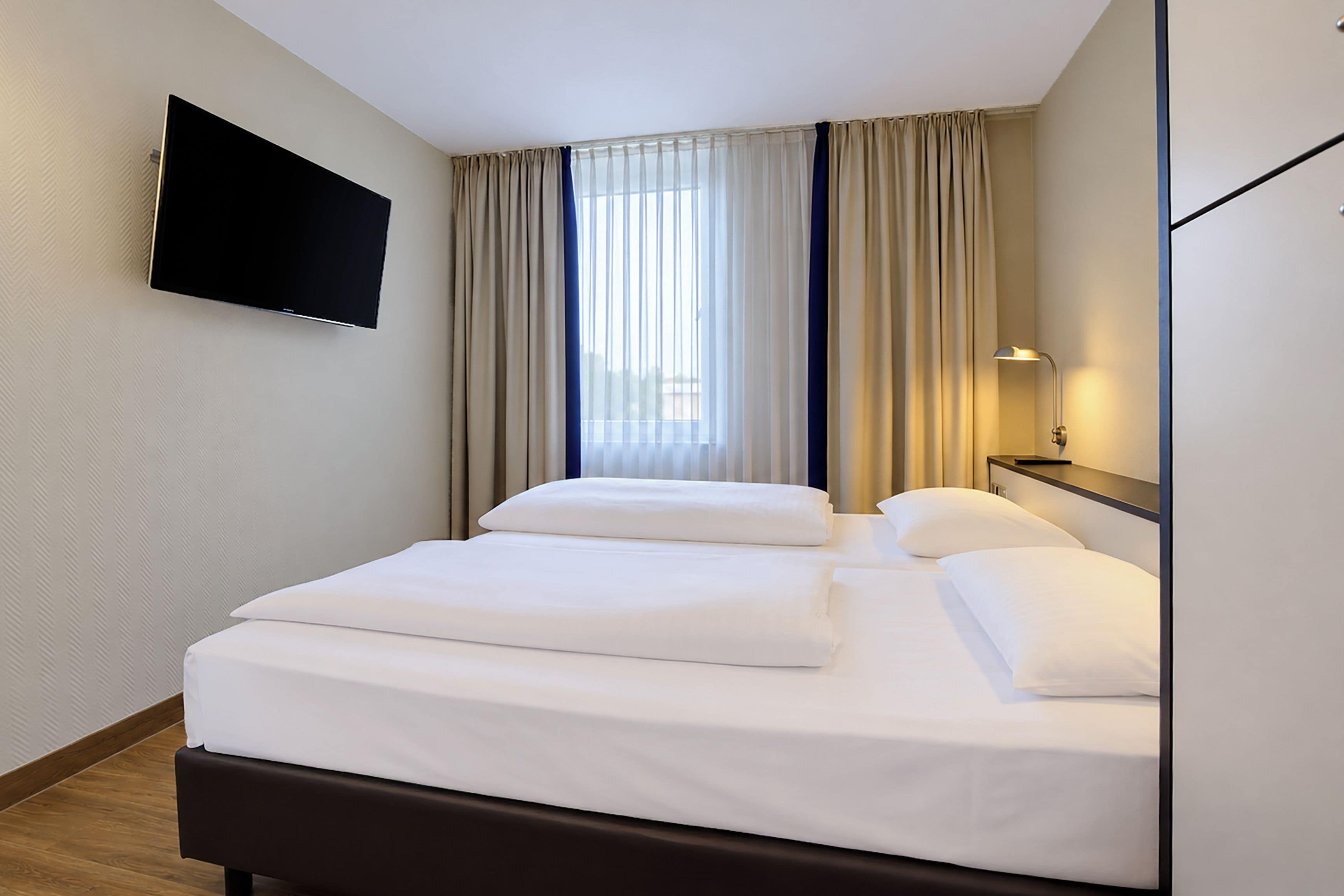Una habitación de hotel con una cama, un televisor de pantalla plana montado en la pared y un ventanal con cortinas.