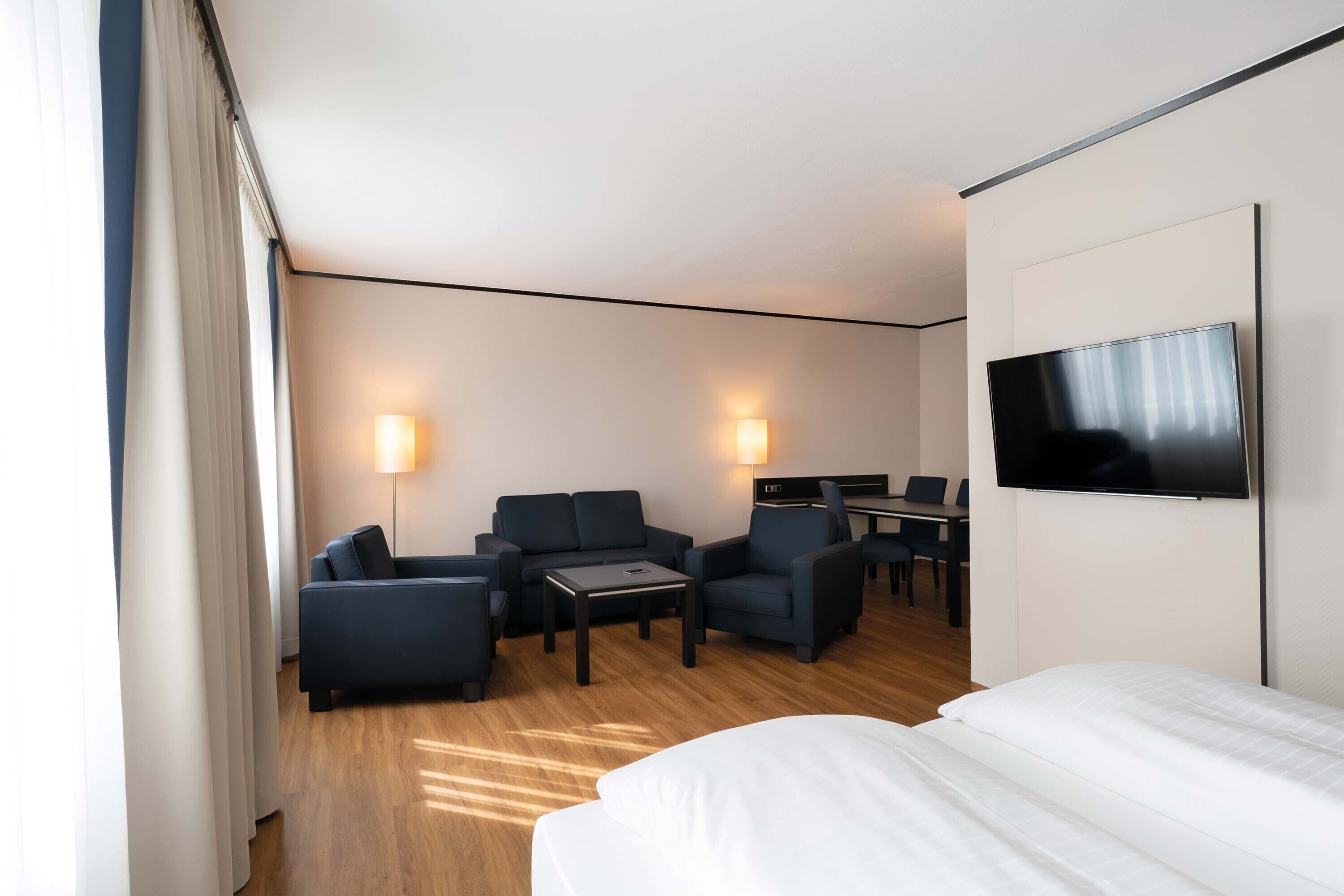 Ein Hotelzimmer mit einem Bett, zwei Sesseln, einem kleinen Tisch, einem Schreibtisch und einem Fernseher.